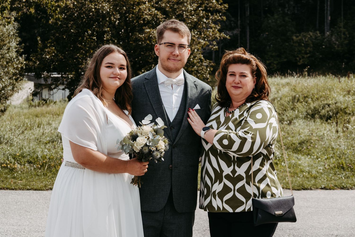 Bride in white dress and groom in suit pose with female guest outdoors at wedding ceremony.