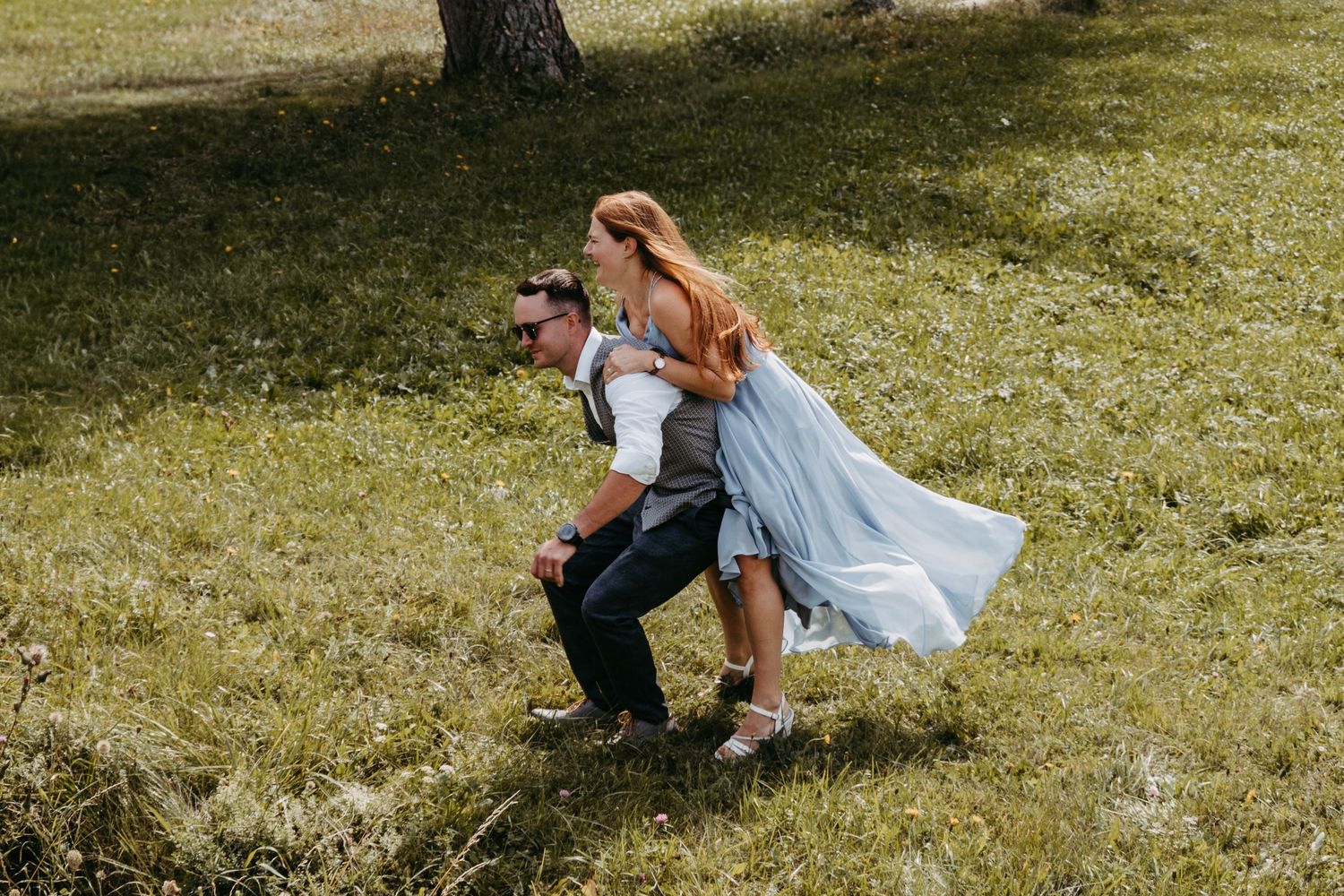 Couple in wedding attire playing outdoors on green grass, bride in blue dress jumping on groom's back.