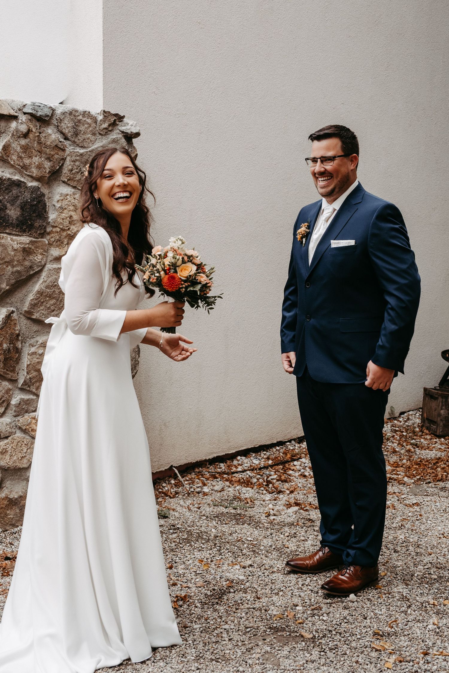 A laughing bride in a white gown holds an autumn bouquet beside her smiling groom in a navy suit.