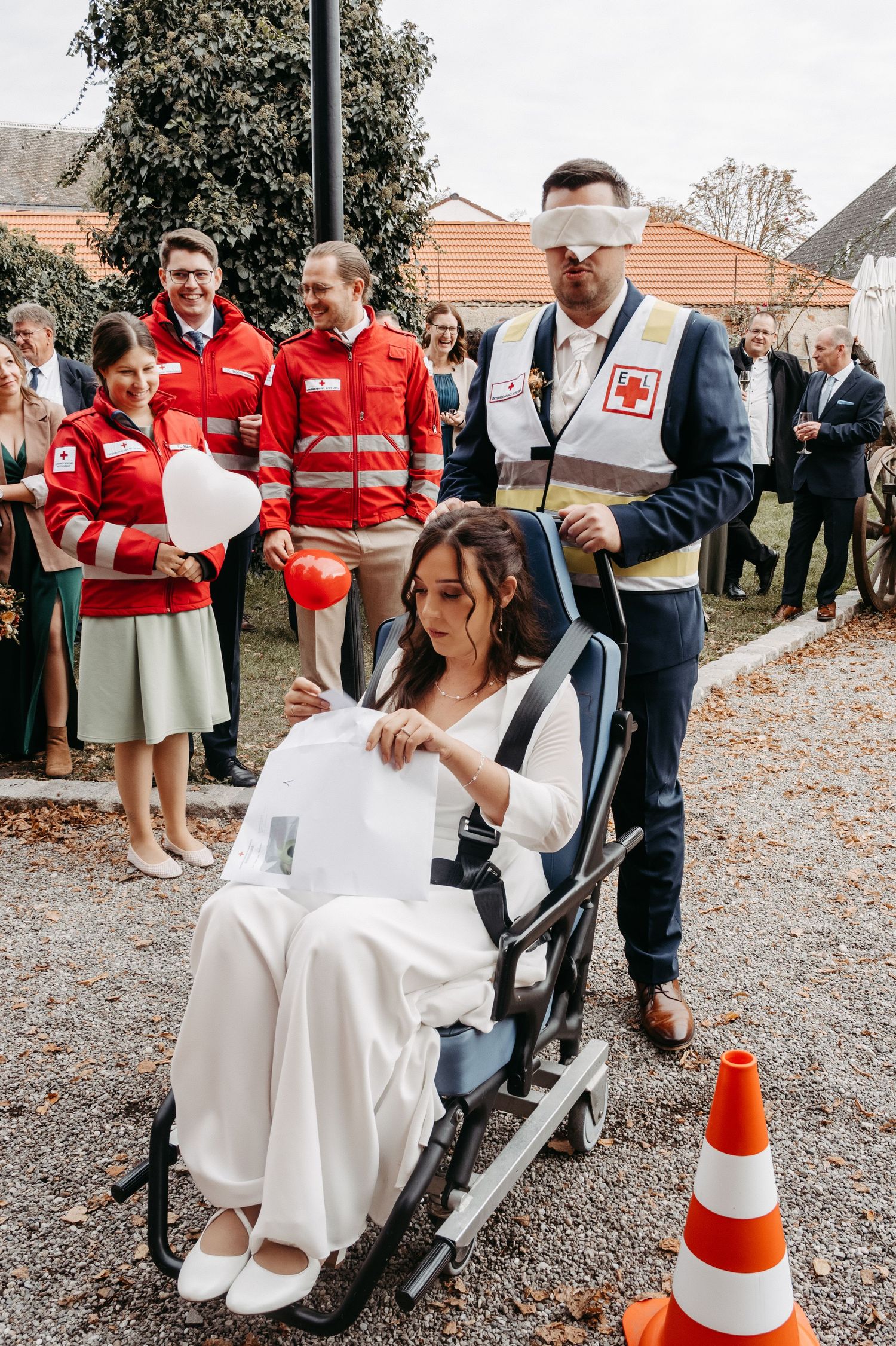 Bride in white dress sits in wheelchair during outdoor wedding game with Red Cross volunteers.