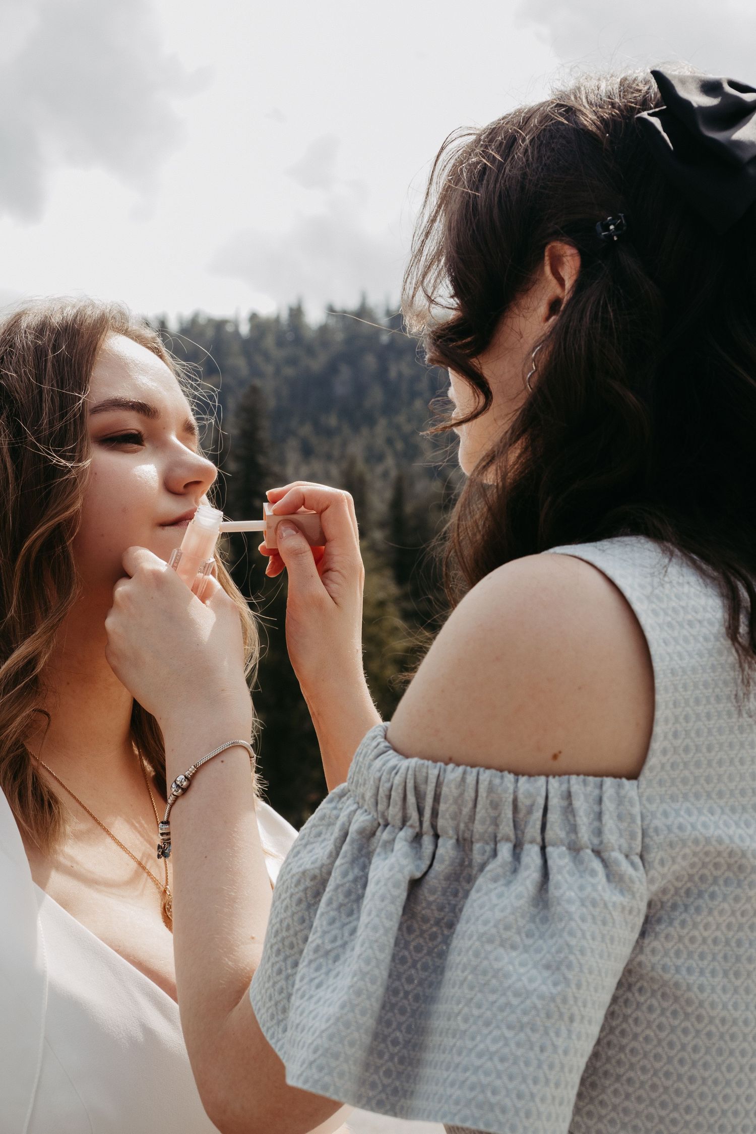 Makeup artist applying lipstick to a young woman outdoors with forest backdrop.