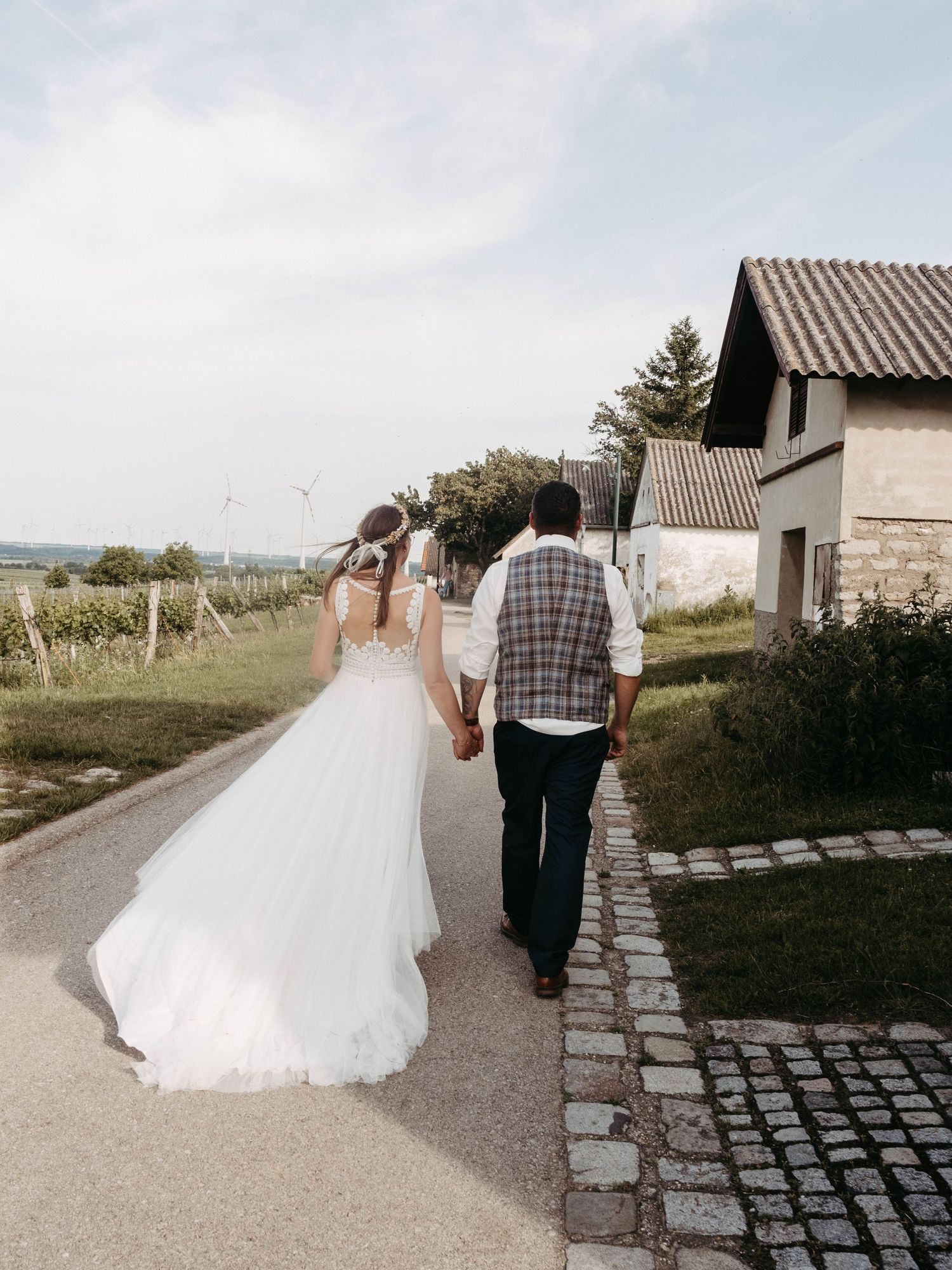 Newlywed couple walking hand in hand down a cobblestone path past rustic buildings and vineyard on their wedding day.