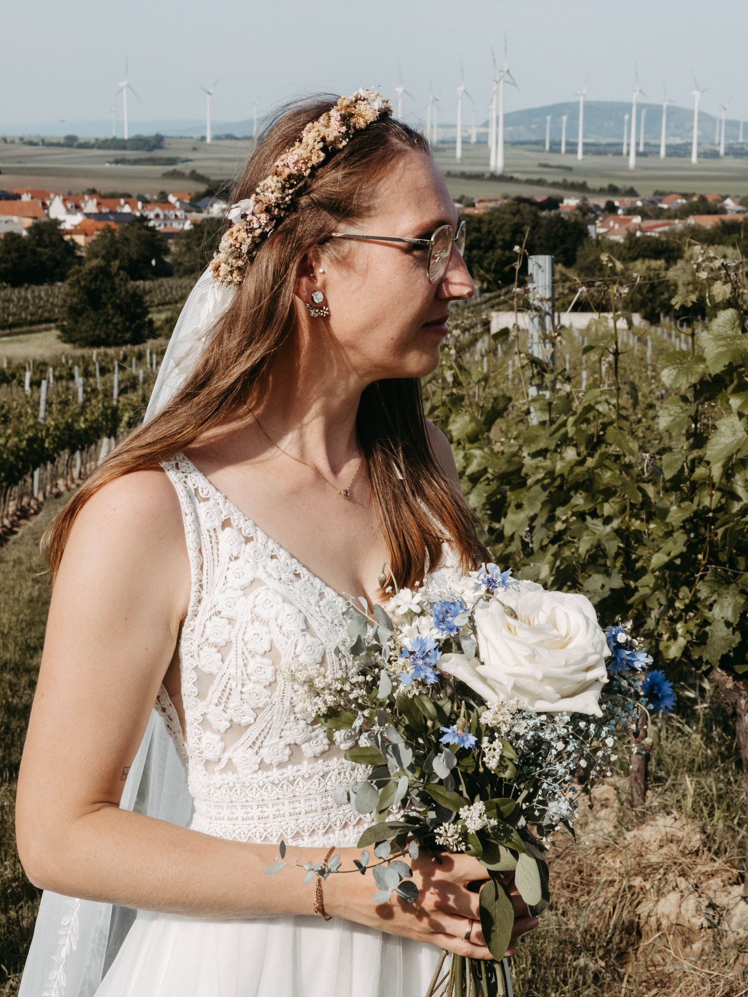 Bride in white lace dress holding blue and white bouquet, wearing floral crown and veil, standing in vineyard on sunny day.