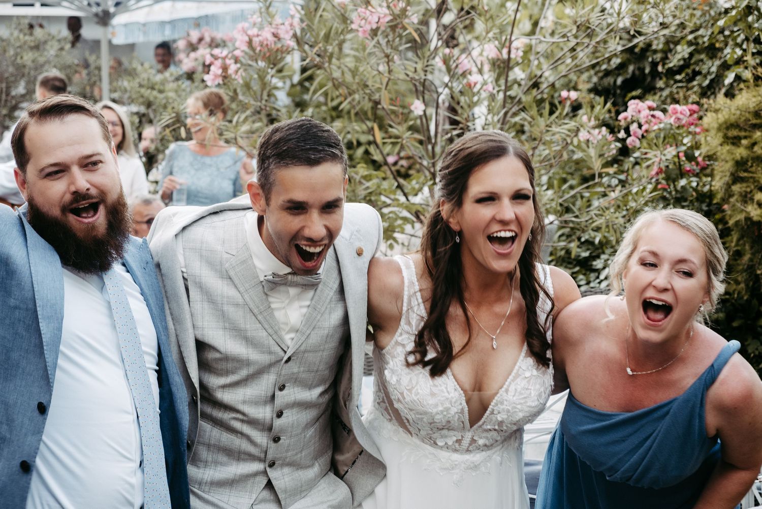 Joyful wedding group photo with bride in white gown and guests laughing outdoors among pink blooming flowers.