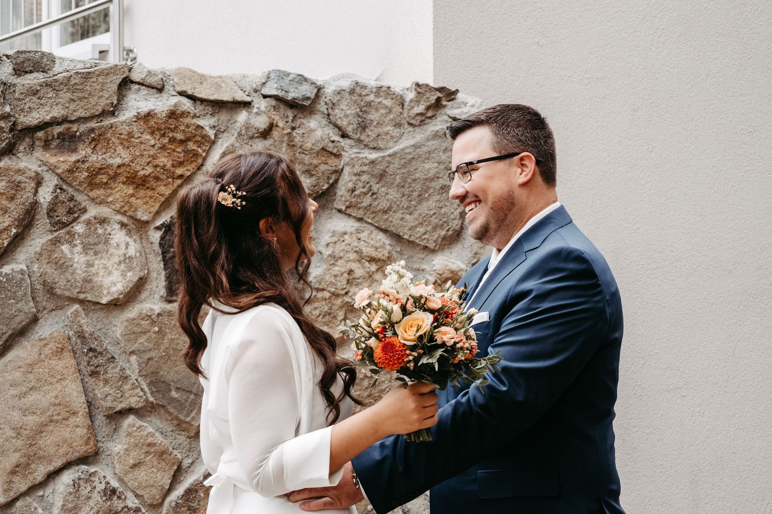 A smiling groom in a navy suit presents a bouquet to his bride outside a stone wall.