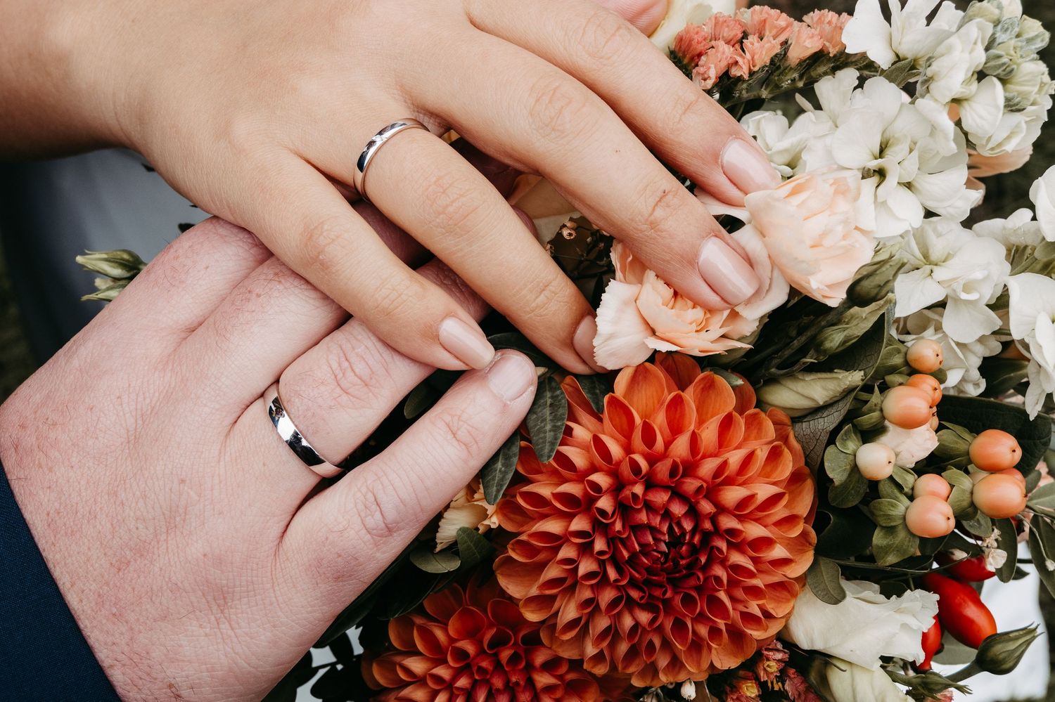 A close-up of newlyweds hands wearing wedding rings resting on a vibrant autumn floral bouquet.
