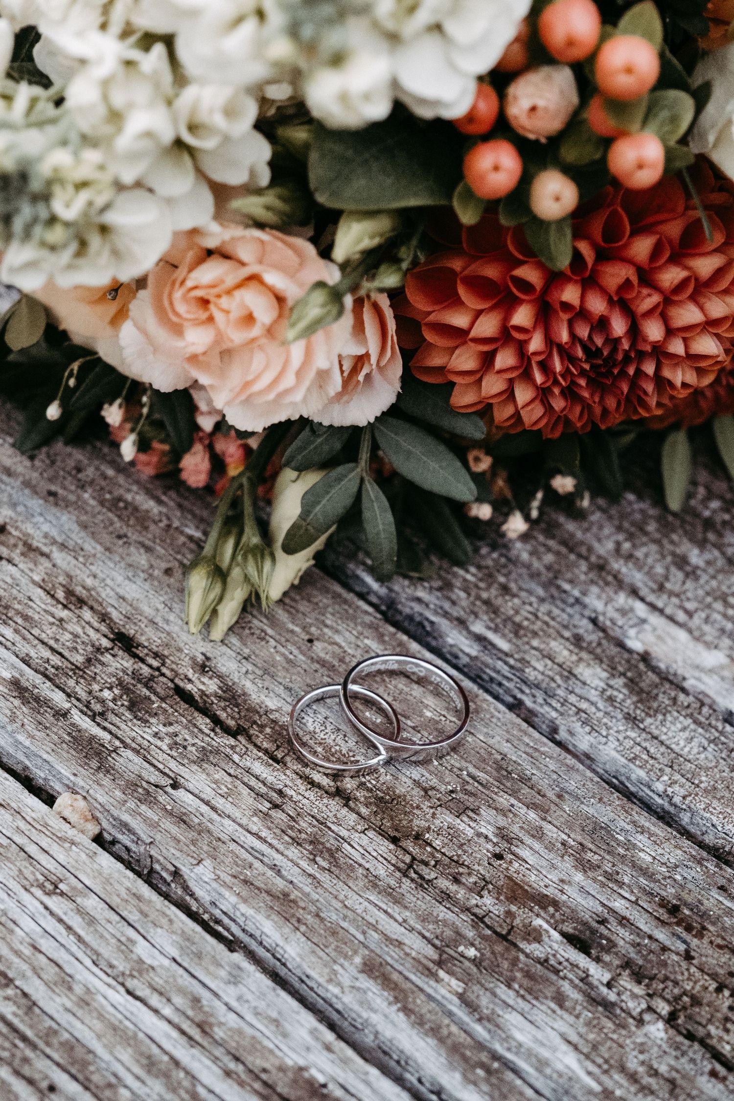 Two silver wedding bands rest on rustic wood beside a bouquet of dahlias, roses, and berries.