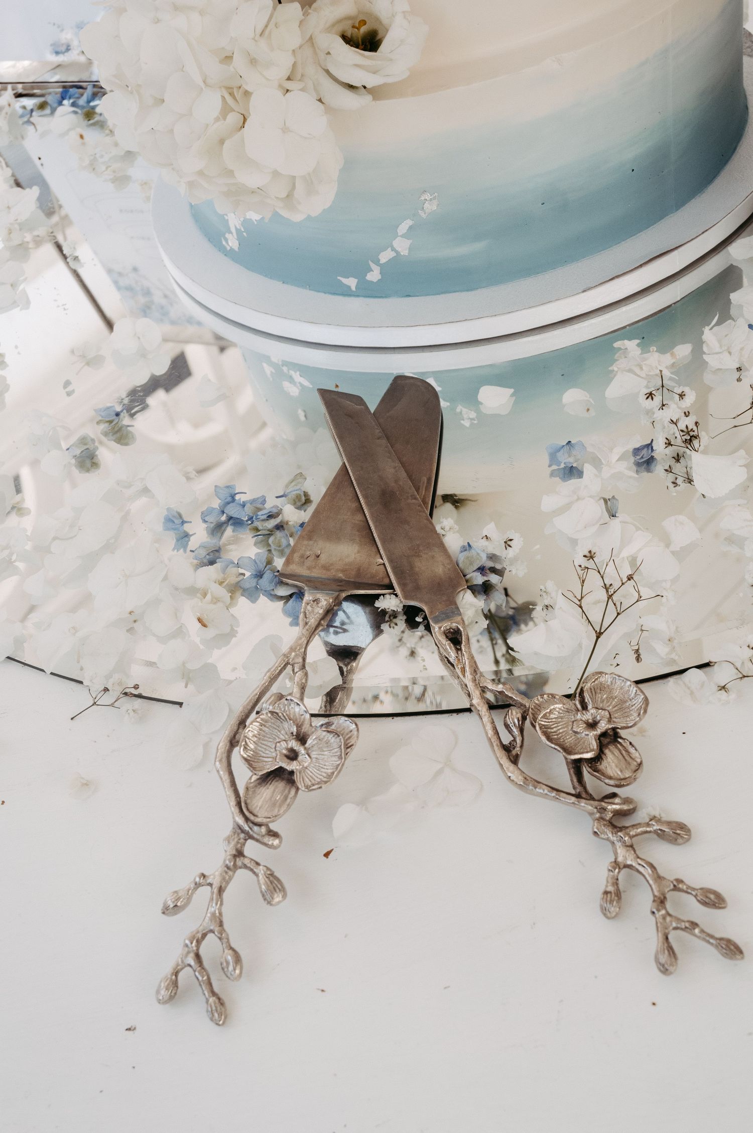 Elegant wedding cake server set with floral details rests on mirror beside blue ombre tiered cake adorned with white flowers.
