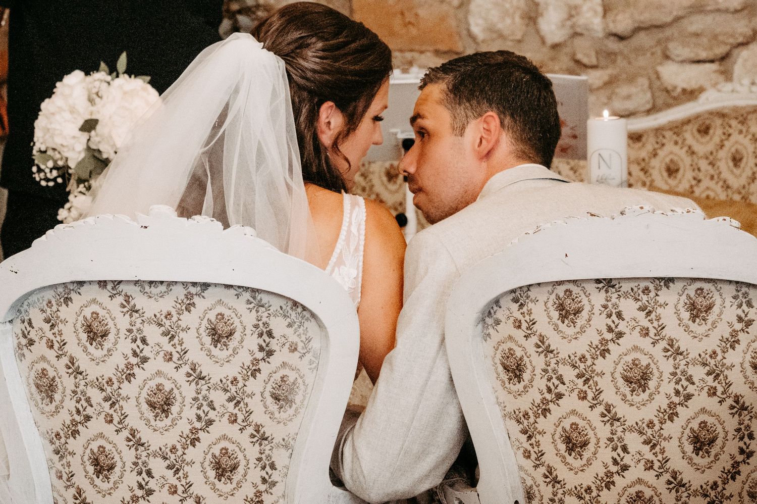 Bride and groom share an intimate moment seated in ornate white chairs during their wedding ceremony.