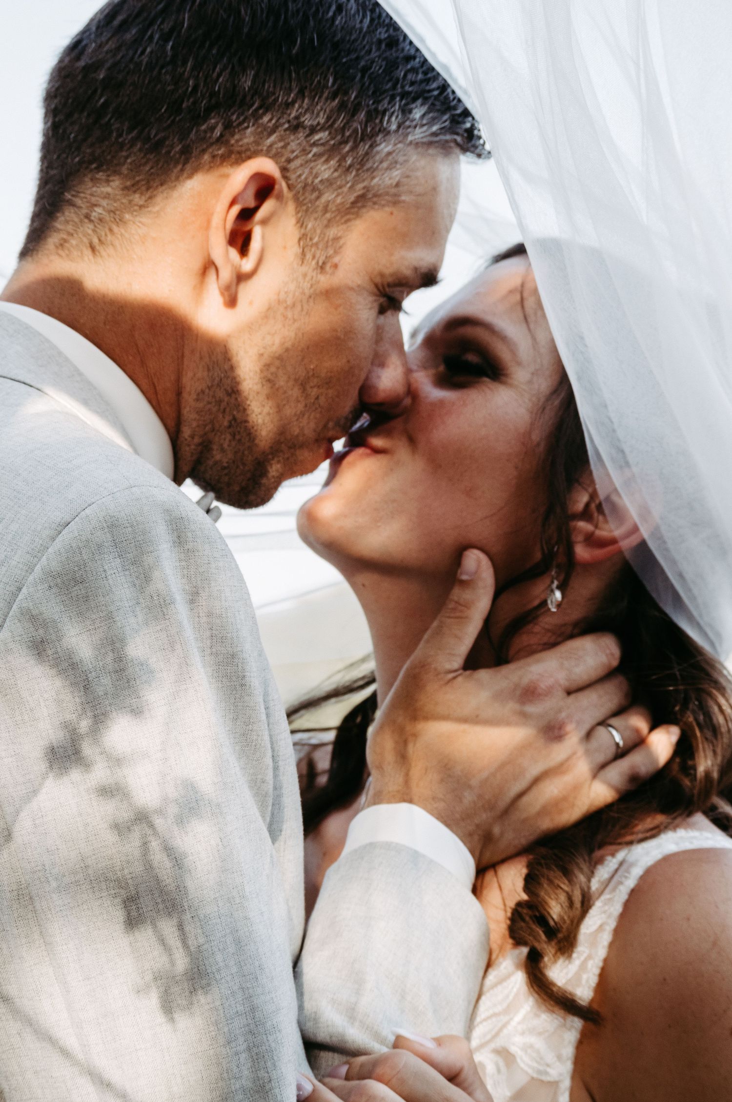 A bride and groom share a romantic kiss under a white veil on their wedding day, with the groom gently holding her face.