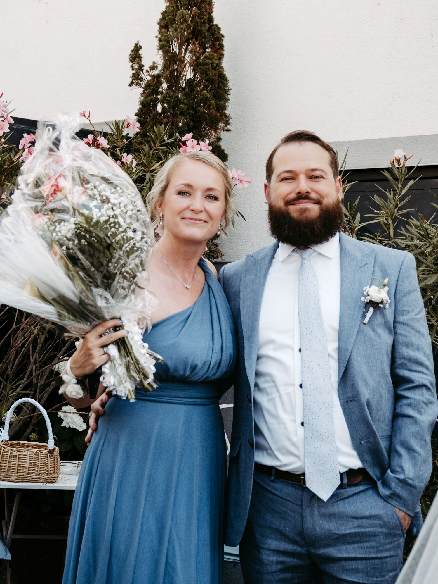 Couple in blue attire at wedding, woman holding bouquet, man with boutonniere, posing outdoors.