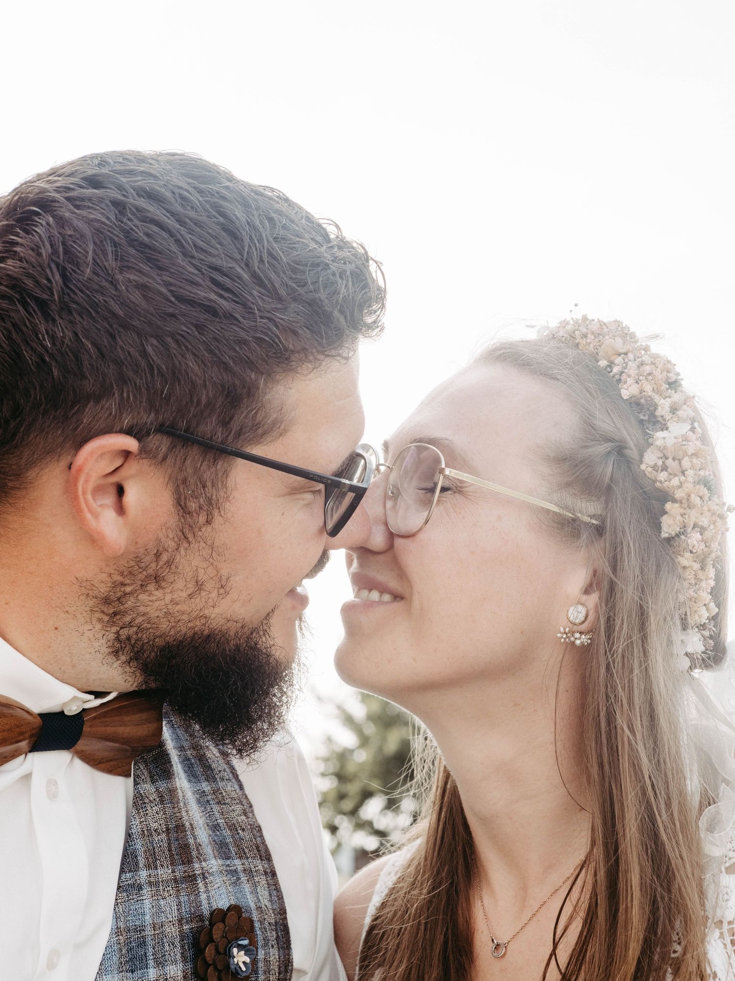 A couple in wedding attire, both wearing glasses, share a romantic nose-to-nose moment, bride wearing a floral crown.