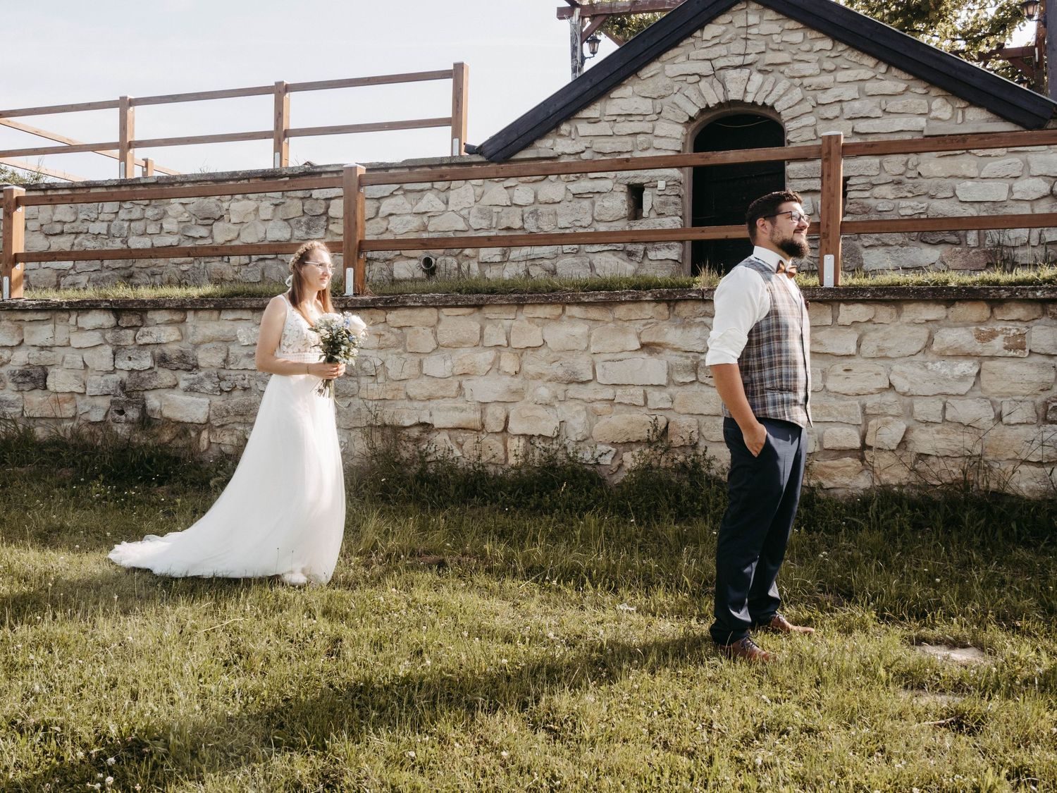 Bride in white gown holds bouquet while groom in vest stands on green lawn beside rustic stone building at outdoor wedding.