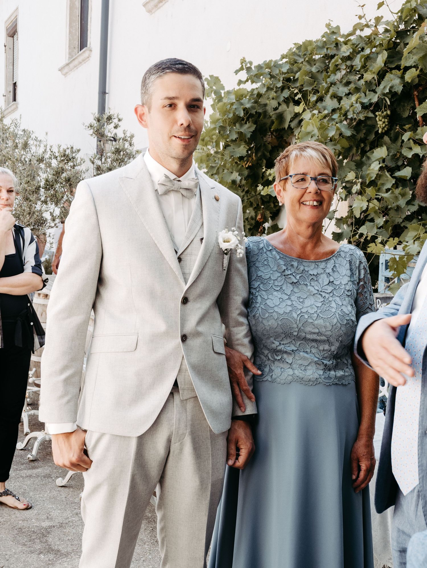 A groom in a light beige suit stands beside a woman in a blue lace dress at an outdoor wedding celebration.