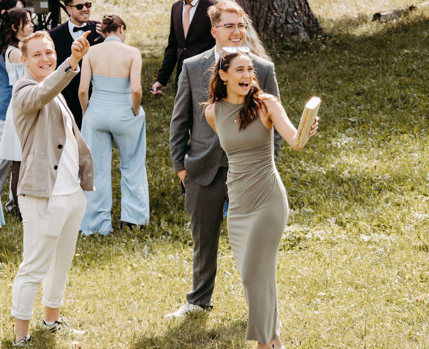 Wedding guests celebrate outdoors on grass, women in elegant dresses holding drinks, smiling joyfully at a summer event.