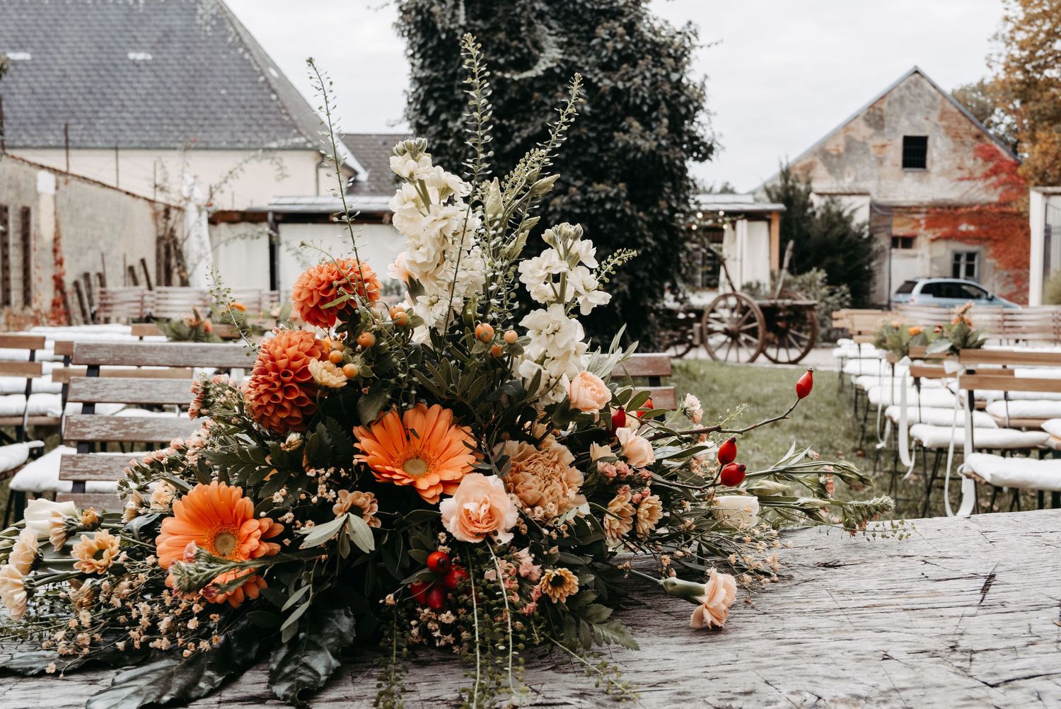 Rustic outdoor wedding ceremony with vibrant orange dahlias and wildflower arrangement on wooden table at a farm venue.