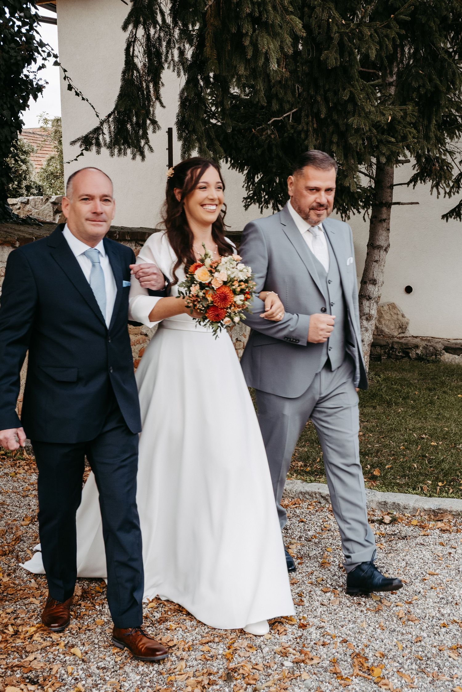 Bride in white gown holding autumn bouquet walks with two men in suits on gravel path.