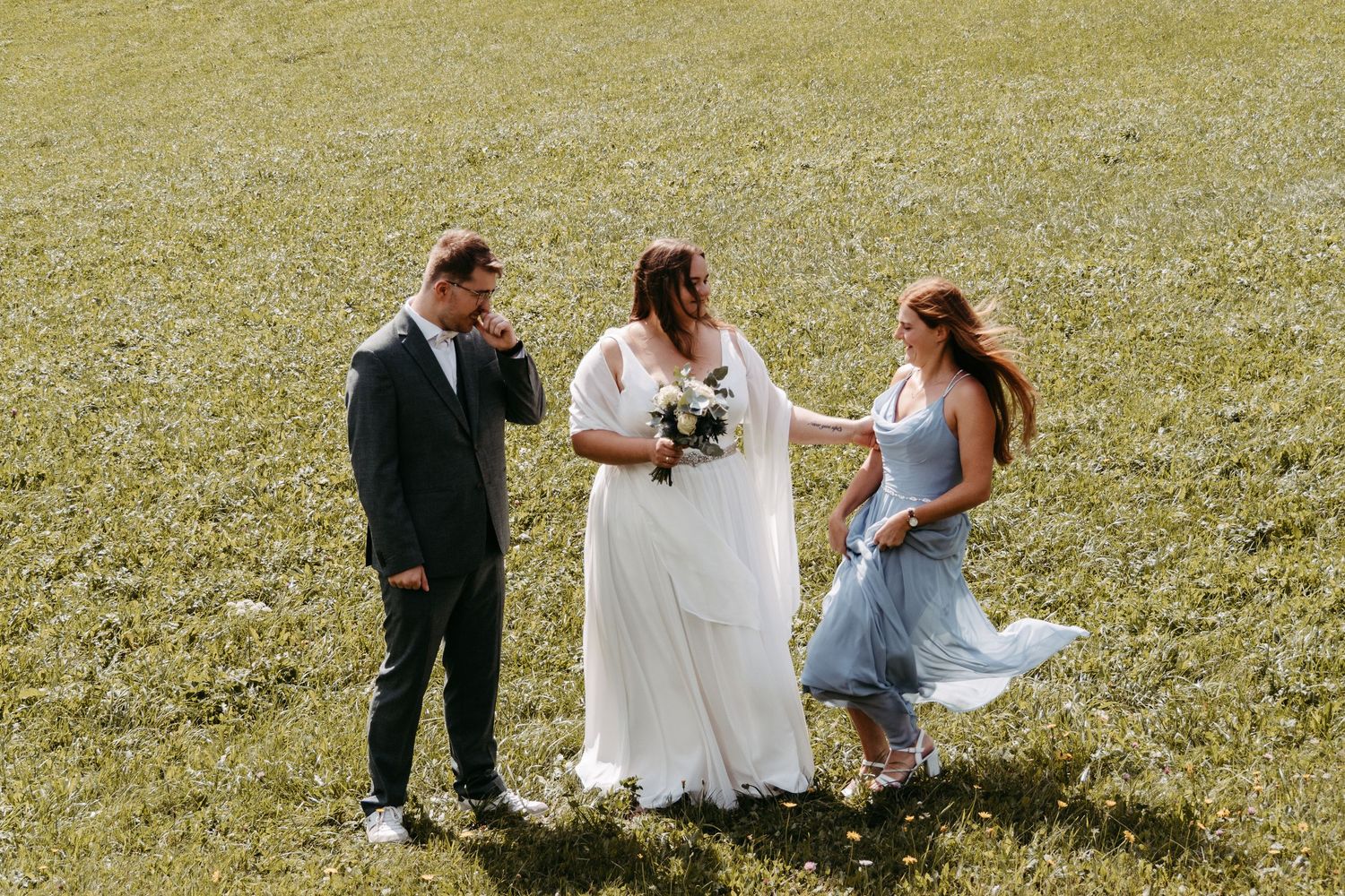 Bride in white gown holds bouquet with groom in suit and bridesmaid in blue dress on green field.