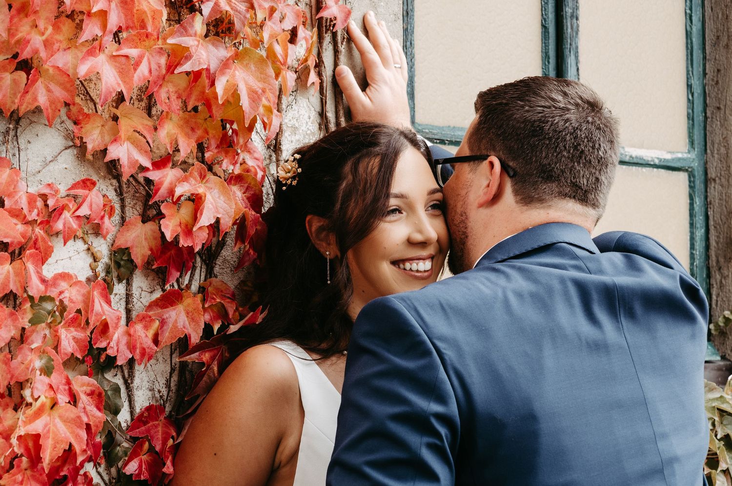 Groom tenderly touching brides hair as she smiles against a vibrant red autumn ivy covered wall.