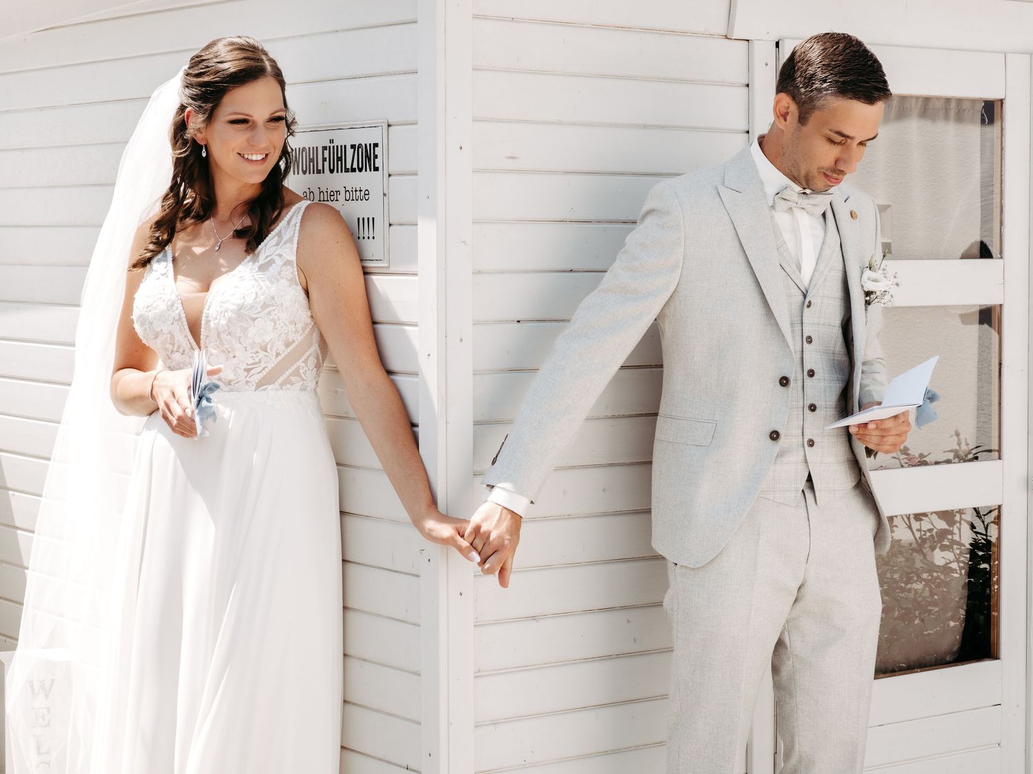 Bride in lace gown and groom in grey suit hold hands around a white building corner, exchanging vows privately.