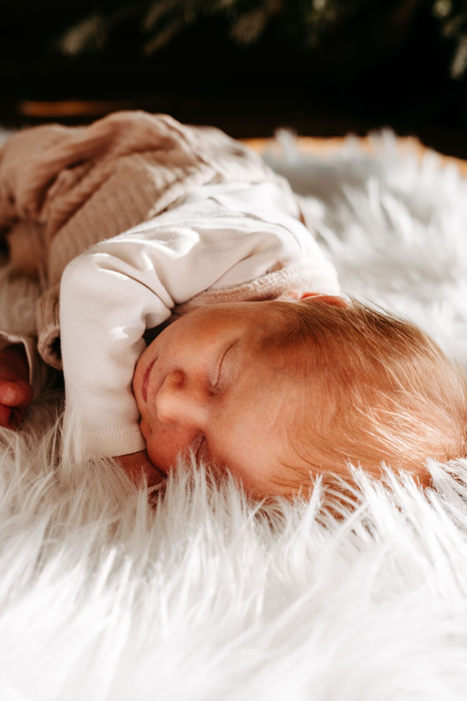 A sleeping infant rests peacefully on soft white fur bedding in a gentle side profile view.