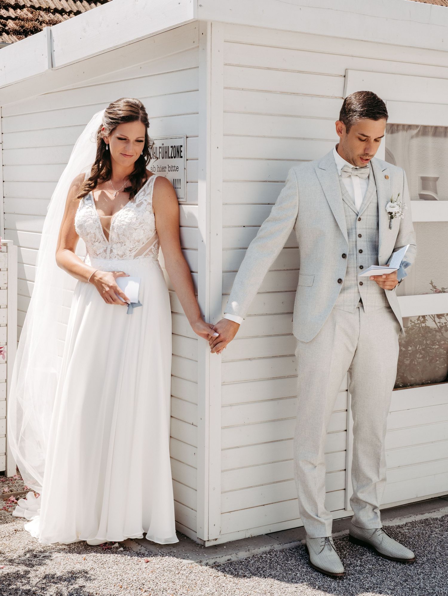 Bride and groom in white attire posing together against white wooden building exterior.