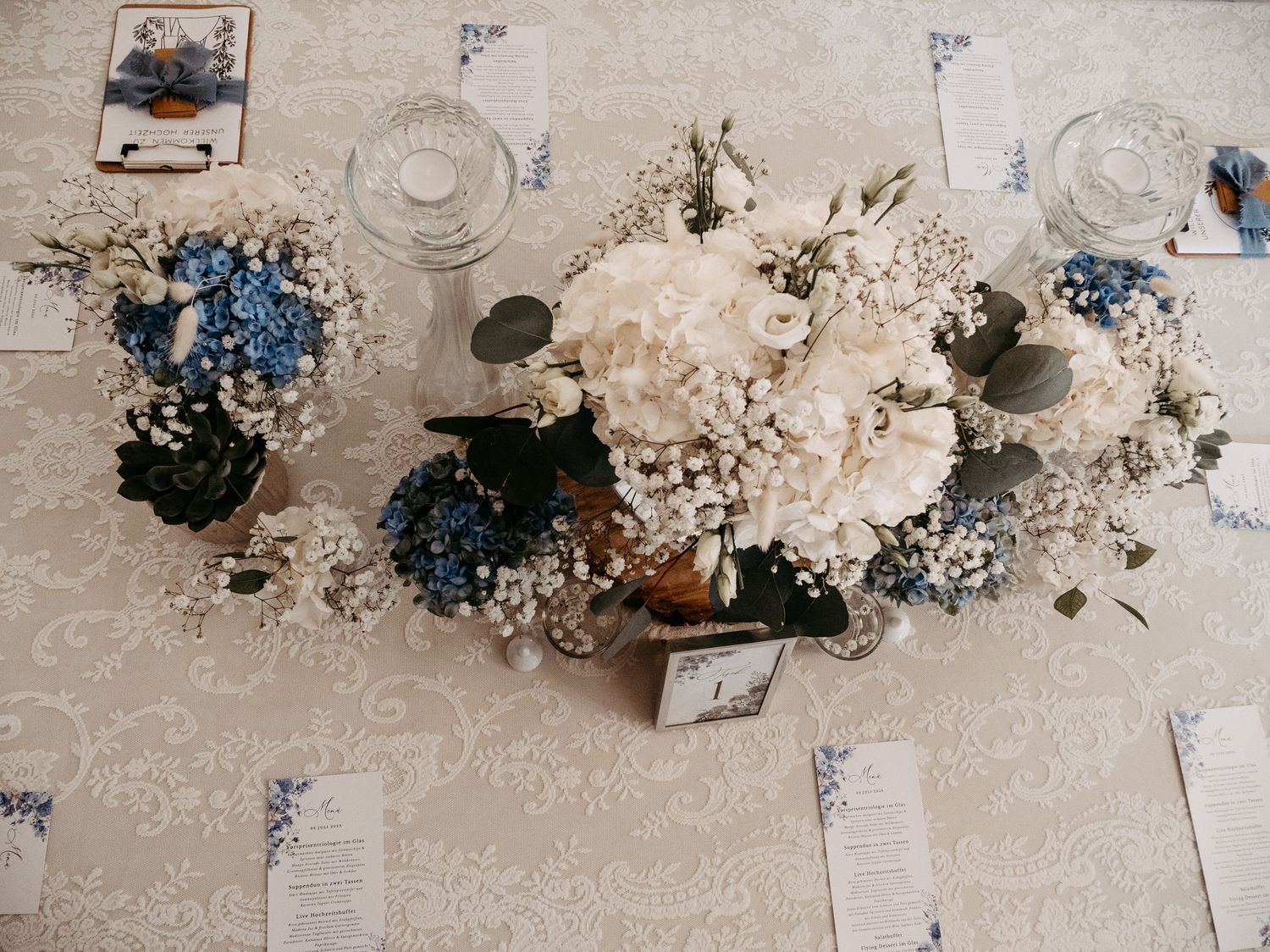 Elegant wedding table centerpiece with white hydrangeas, blue flowers, and baby's breath on a cream lace tablecloth.