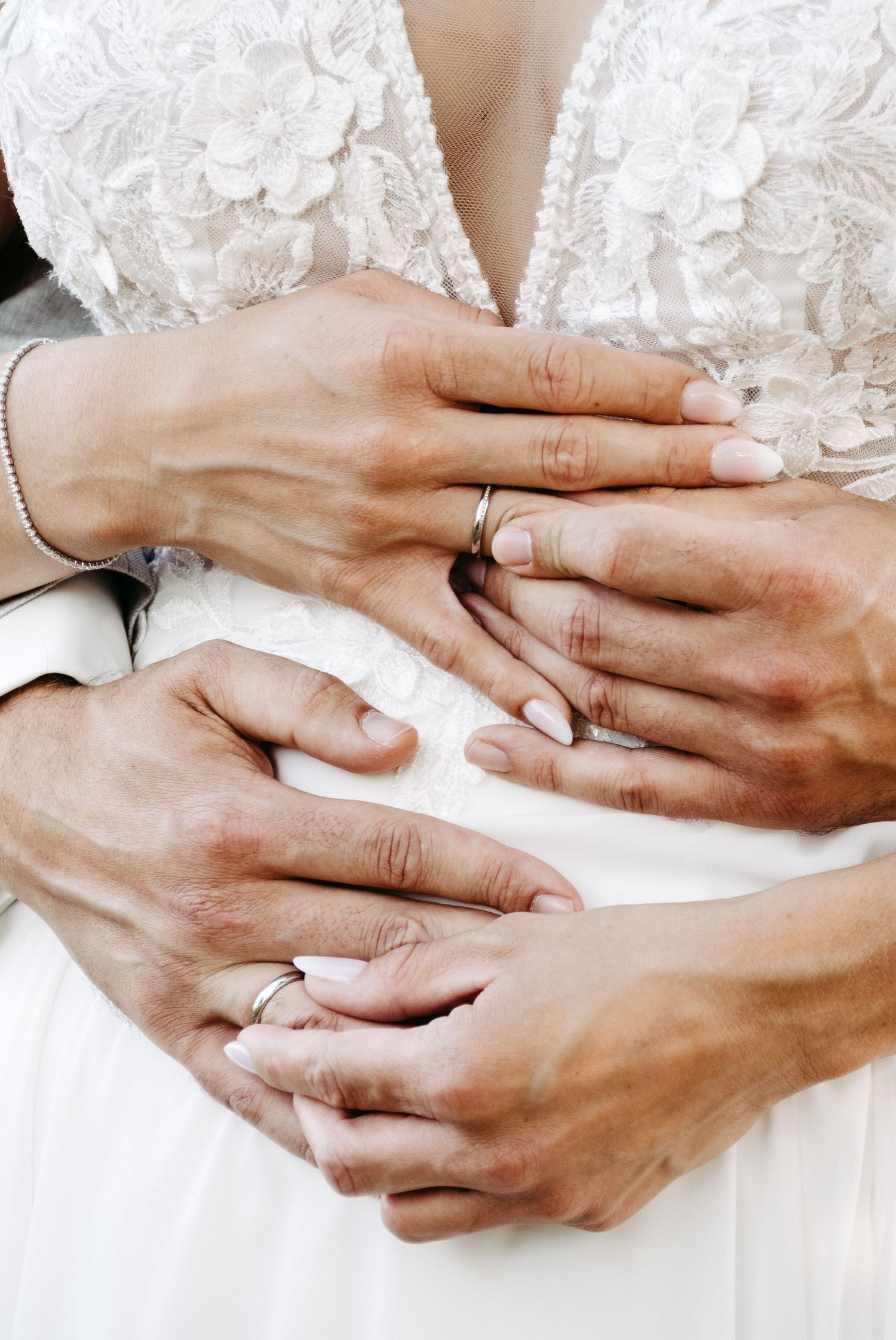 Newlywed couple displaying wedding rings on their hands, bride in white lace dress with groom embracing her waist.