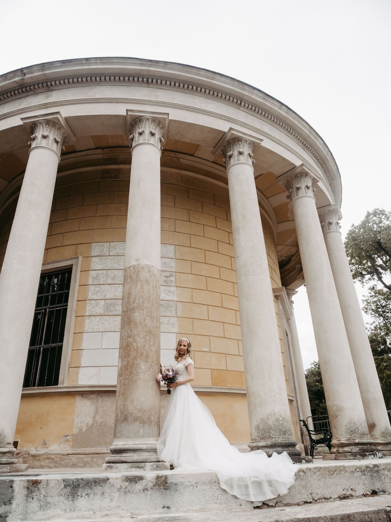 Bride in white gown poses on steps of neoclassical rotunda with tall columns at elegant outdoor wedding venue.