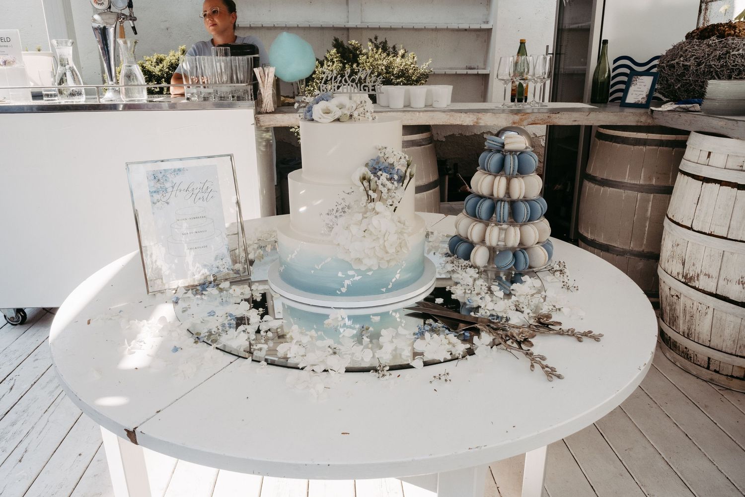 Elegant white and blue wedding cake on a round table decorated with flowers and macarons in a coastal venue.