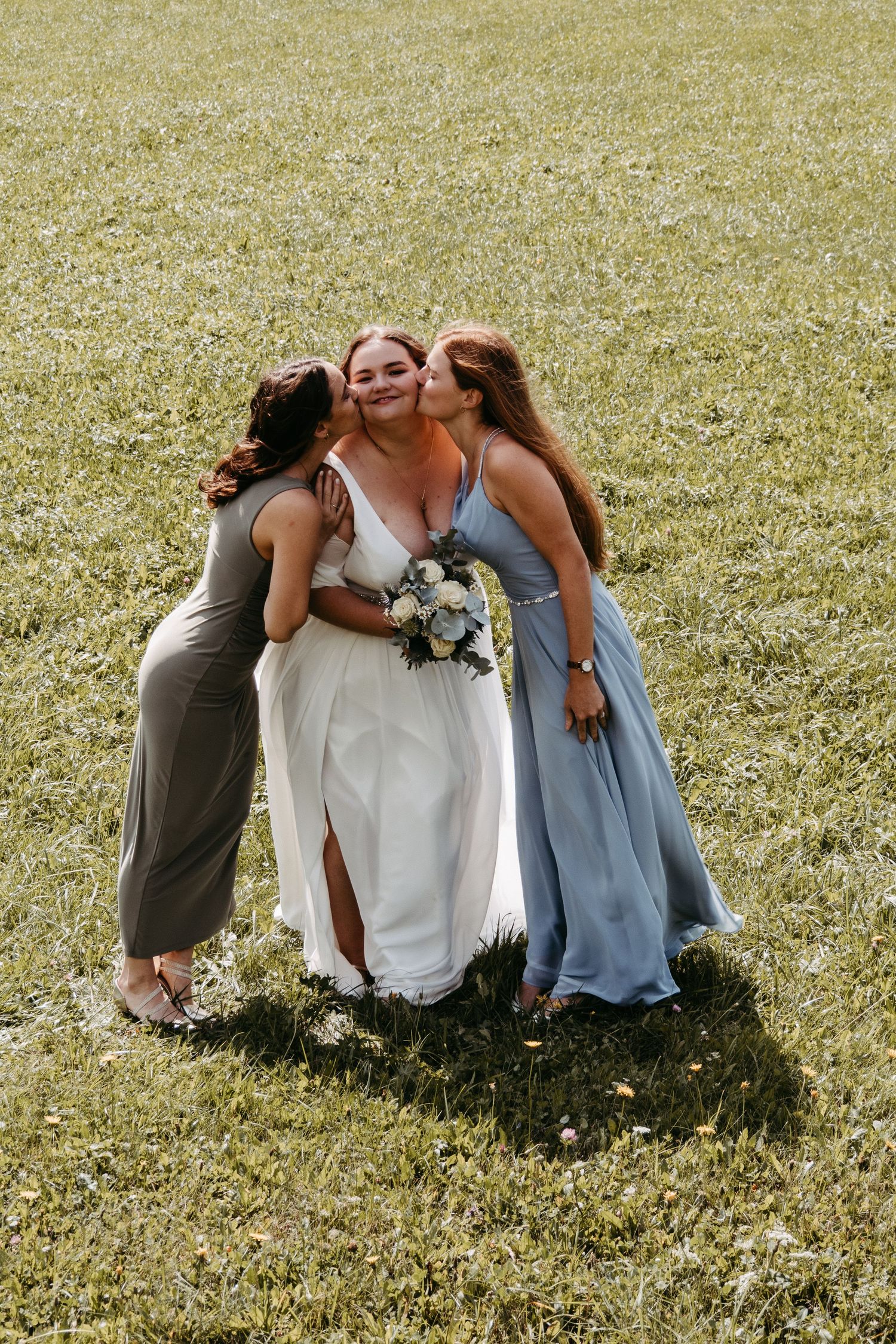 Two bridesmaids kiss smiling bride holding bouquet on green meadow at outdoor wedding.