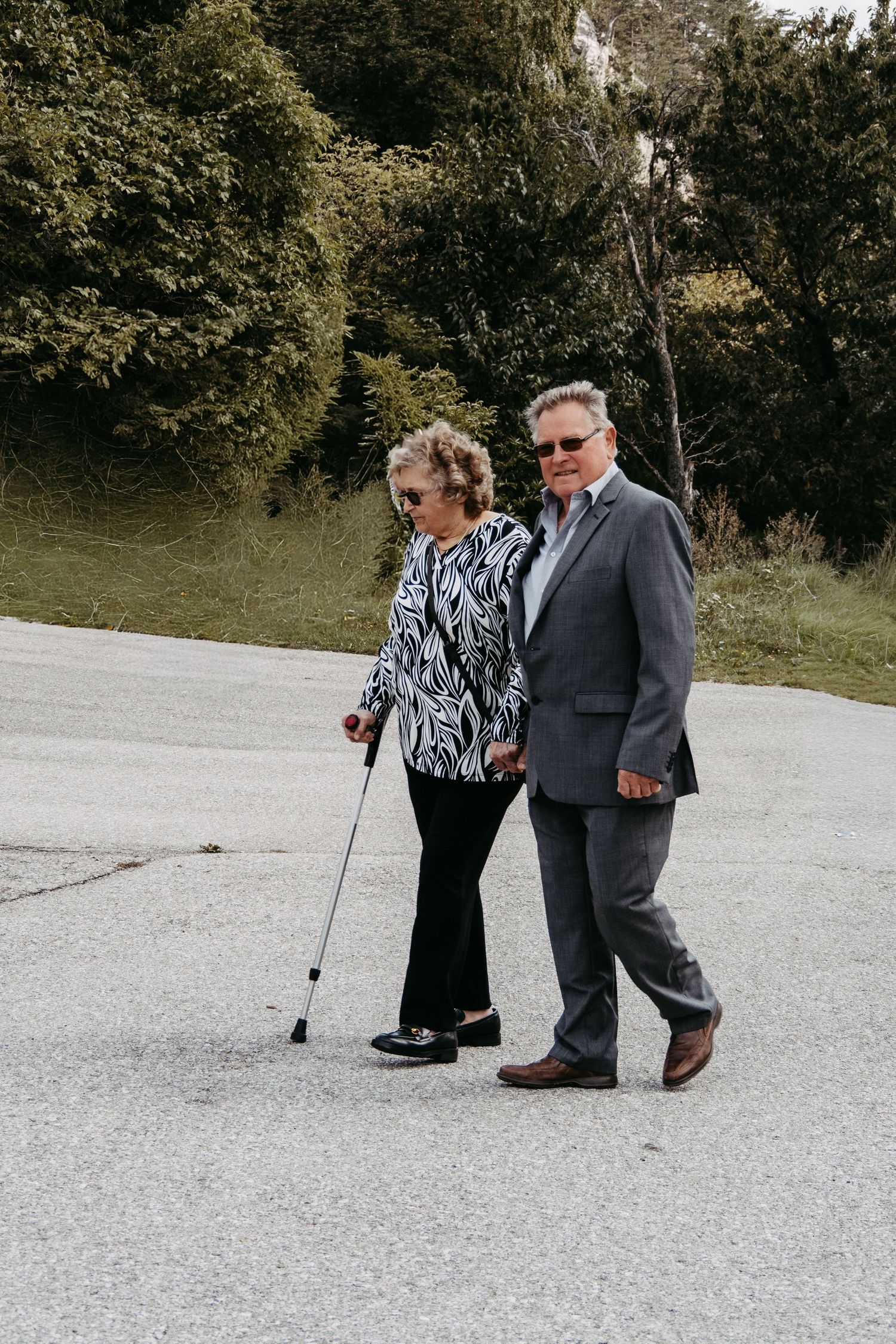 Elderly couple walking outdoors on pavement, woman using a cane, man in gray suit, with green trees in background.