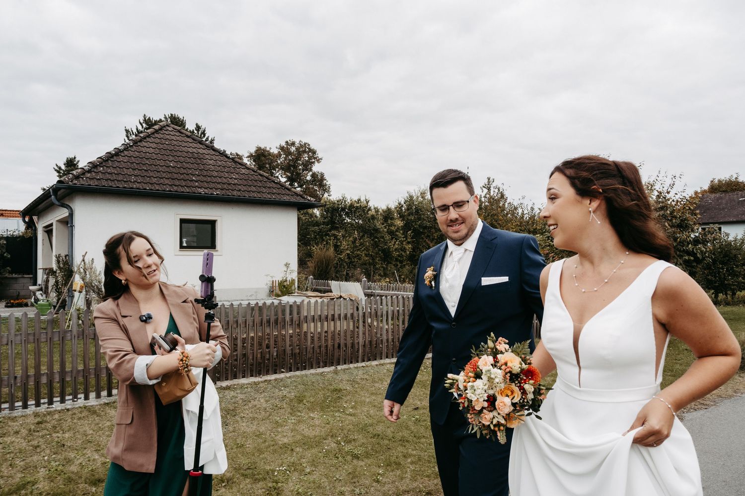Bride and groom laughing with wedding guest holding broom in outdoor setting.