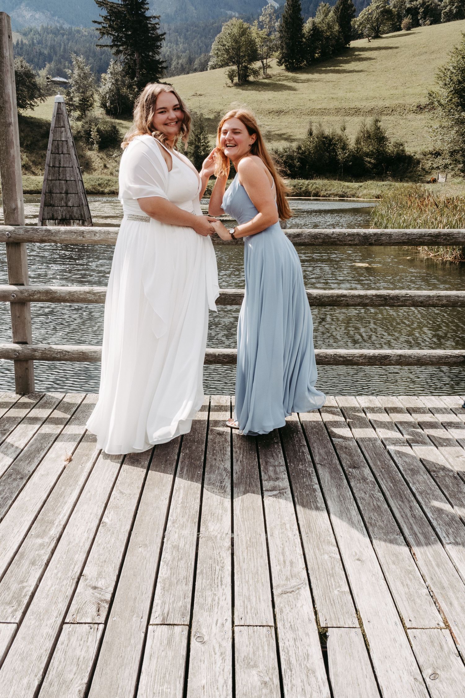 Bride in white gown and bridesmaid in blue dress pose on wooden dock by mountain lake.