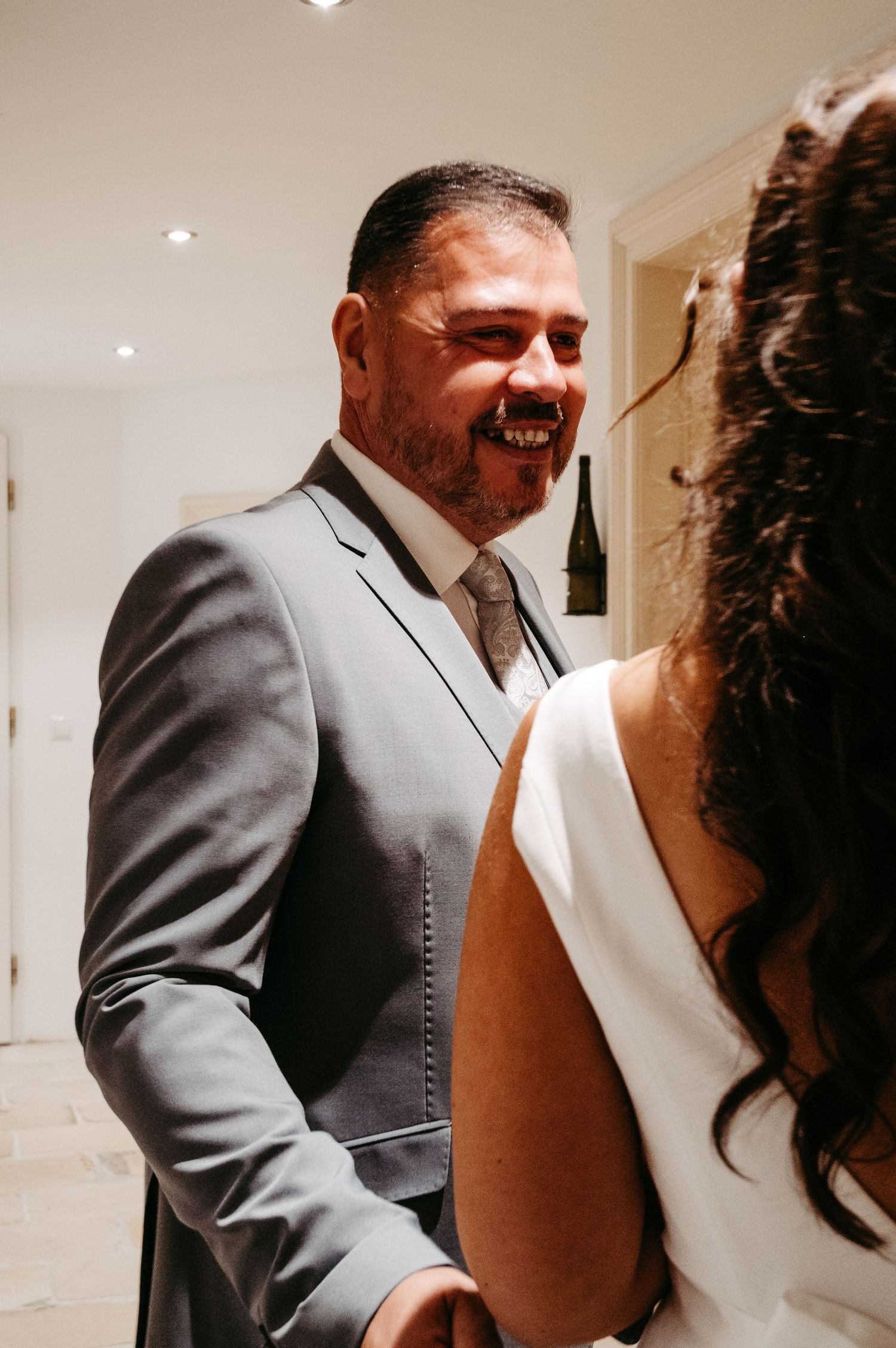 Smiling man in gray suit laughing with bride in white backless dress at wedding reception.