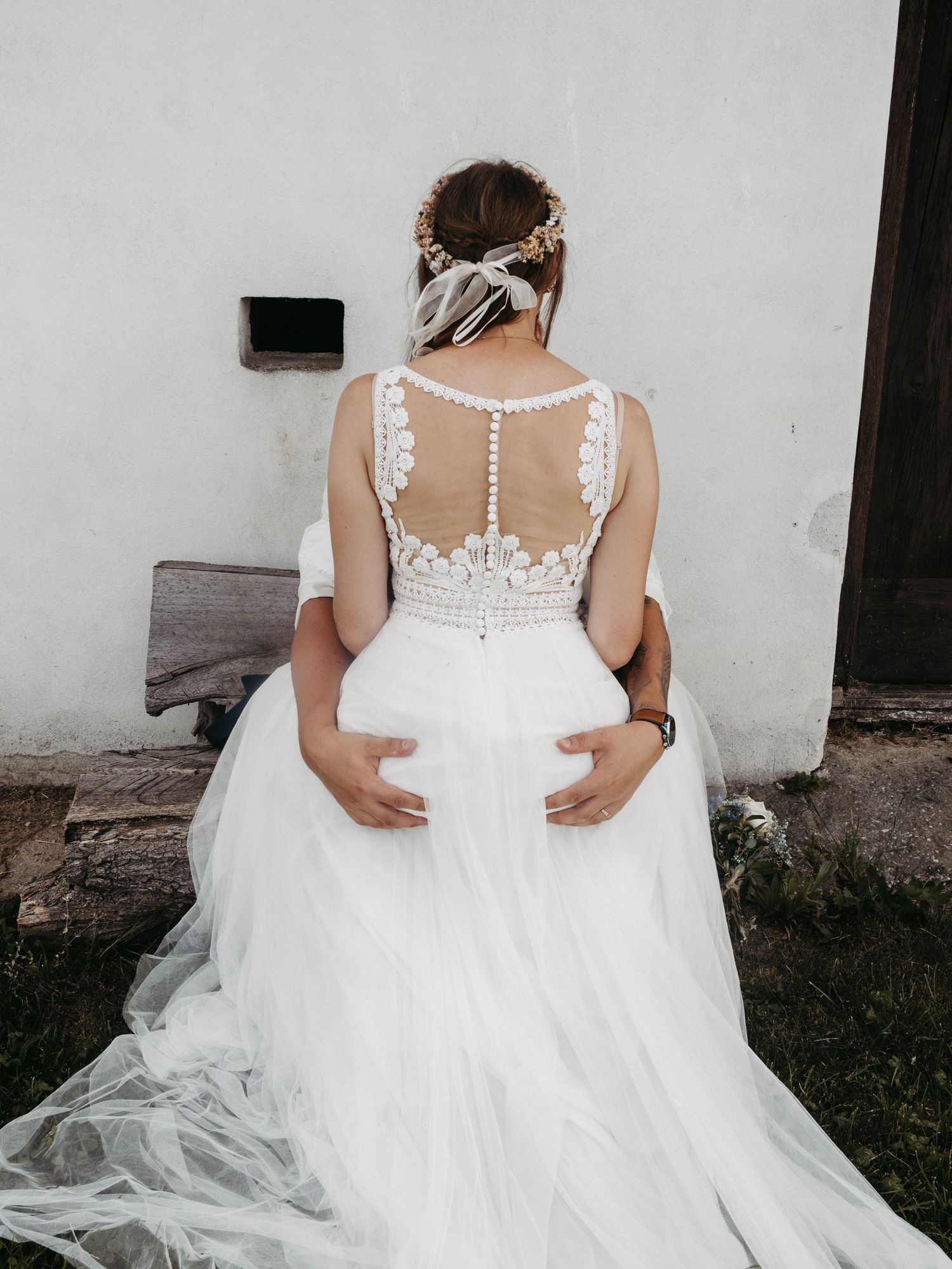 Bride in white lace wedding gown with tulle skirt, viewed from behind, with floral hair accessories outdoors.