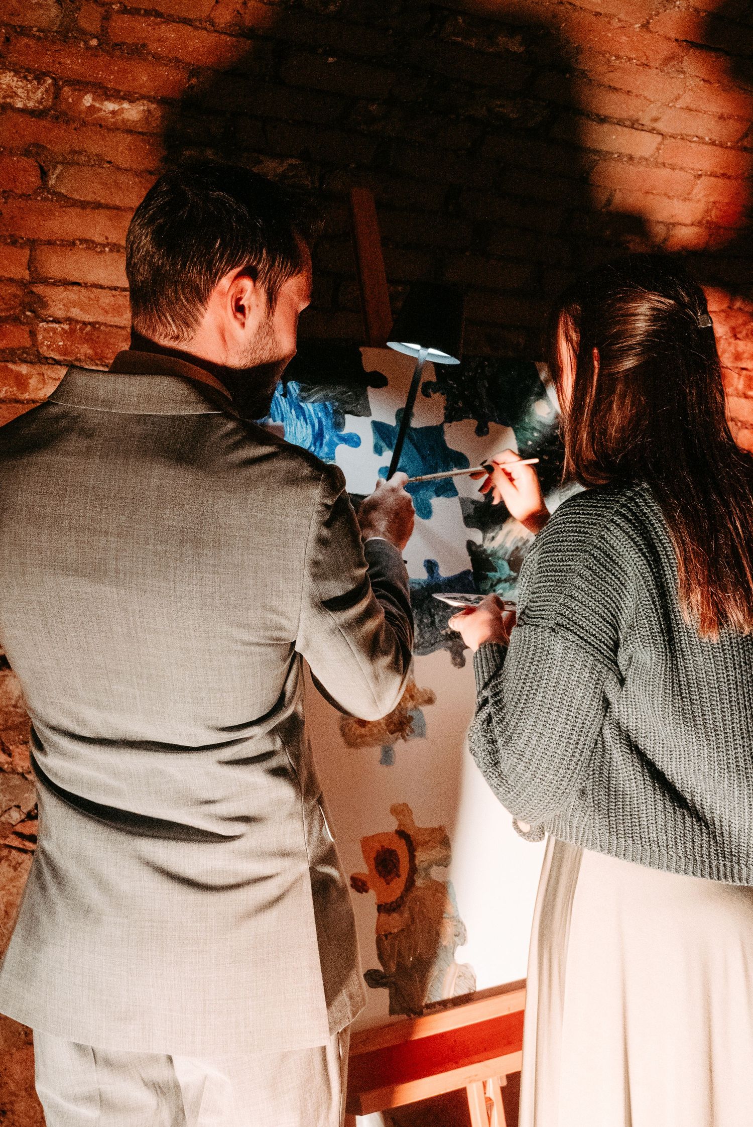 A couple in formal attire works together on a colorful puzzle painting at an easel indoors.