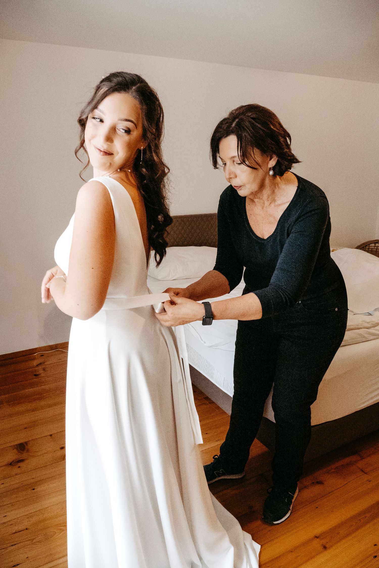 A woman in a white wedding dress gets her gown fastened by a helper in a bedroom.