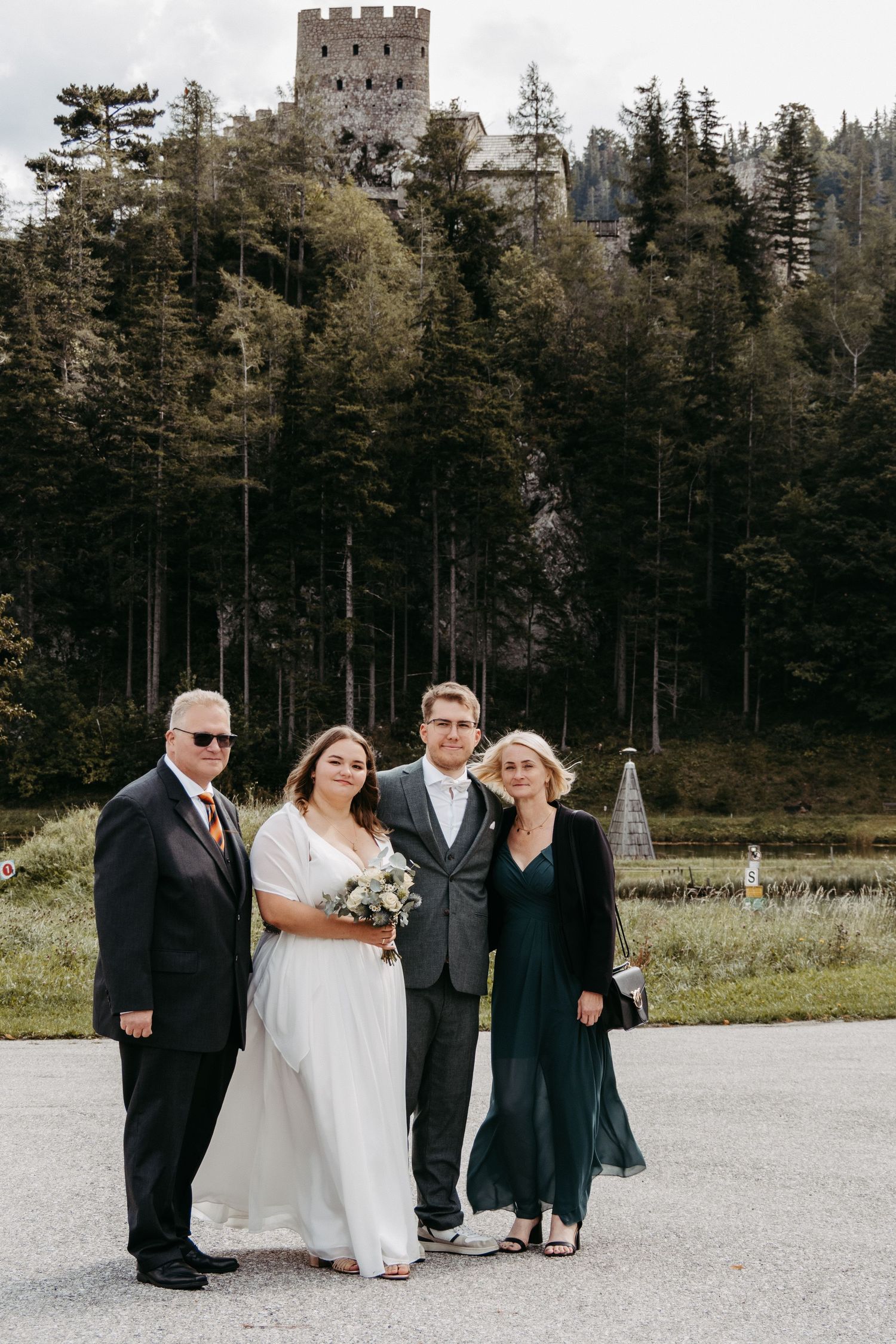 Wedding party of four posing outdoors with a castle and pine forest backdrop on a cloudy day.