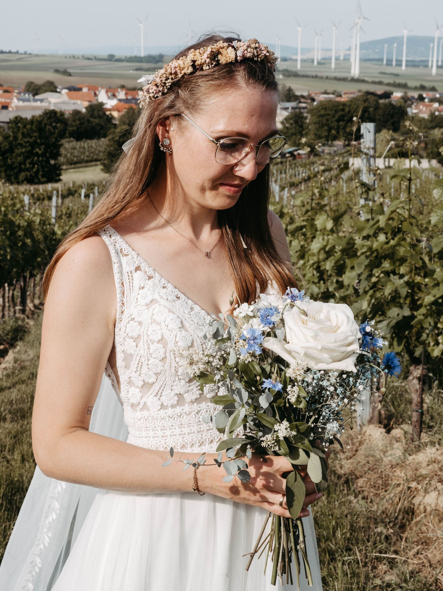 Bride in white lace gown holding blue and white bouquet, wearing floral crown, standing in vineyard with windmills.