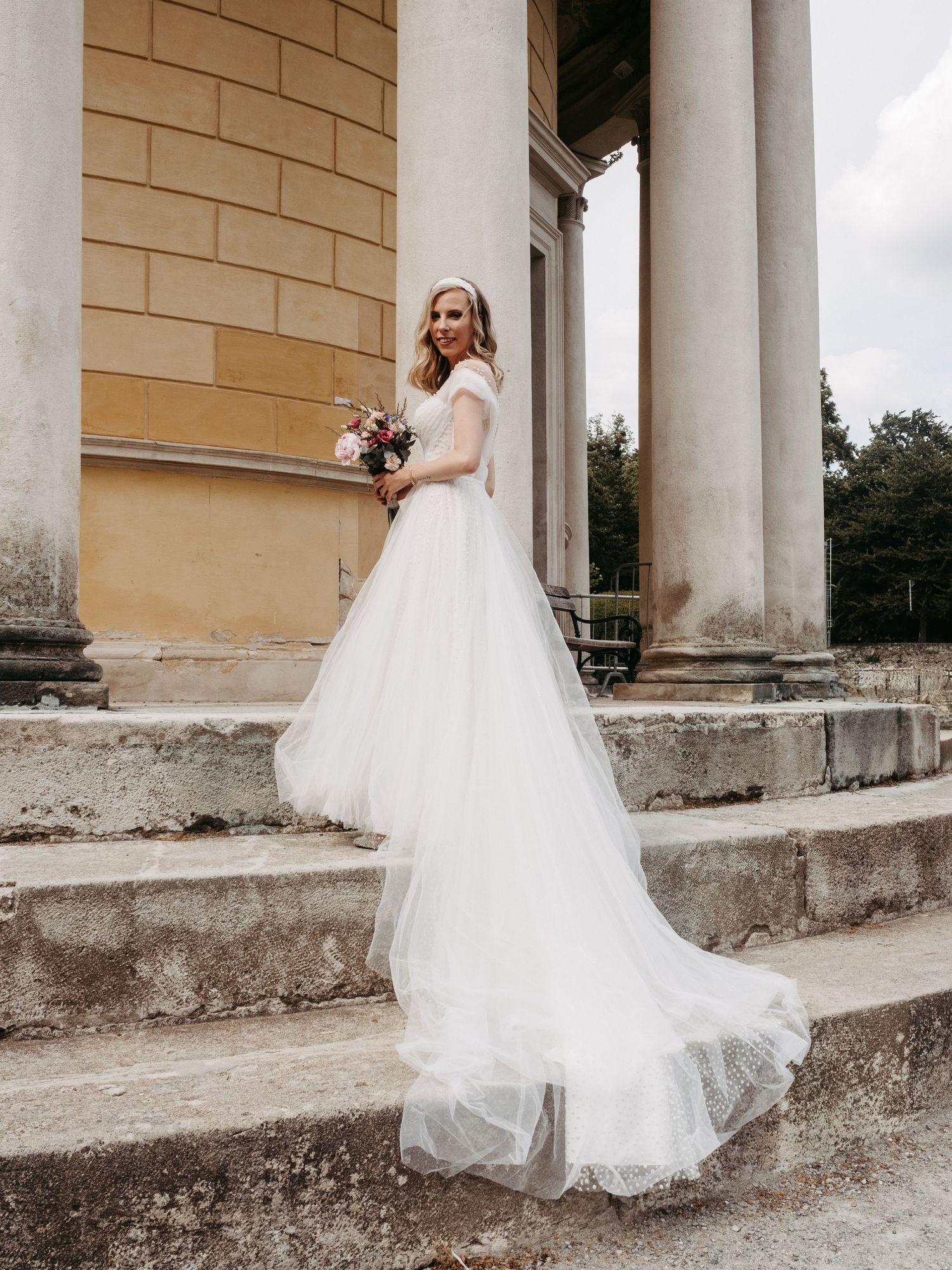 Bride in white tulle wedding gown holding bouquet on stone steps beside classical columns.