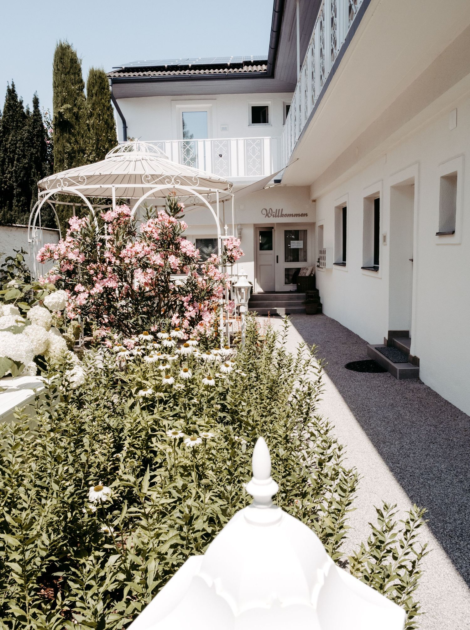 Elegant white hotel exterior with ornate gazebo, blooming pink roses, and lush green garden on a sunny day.