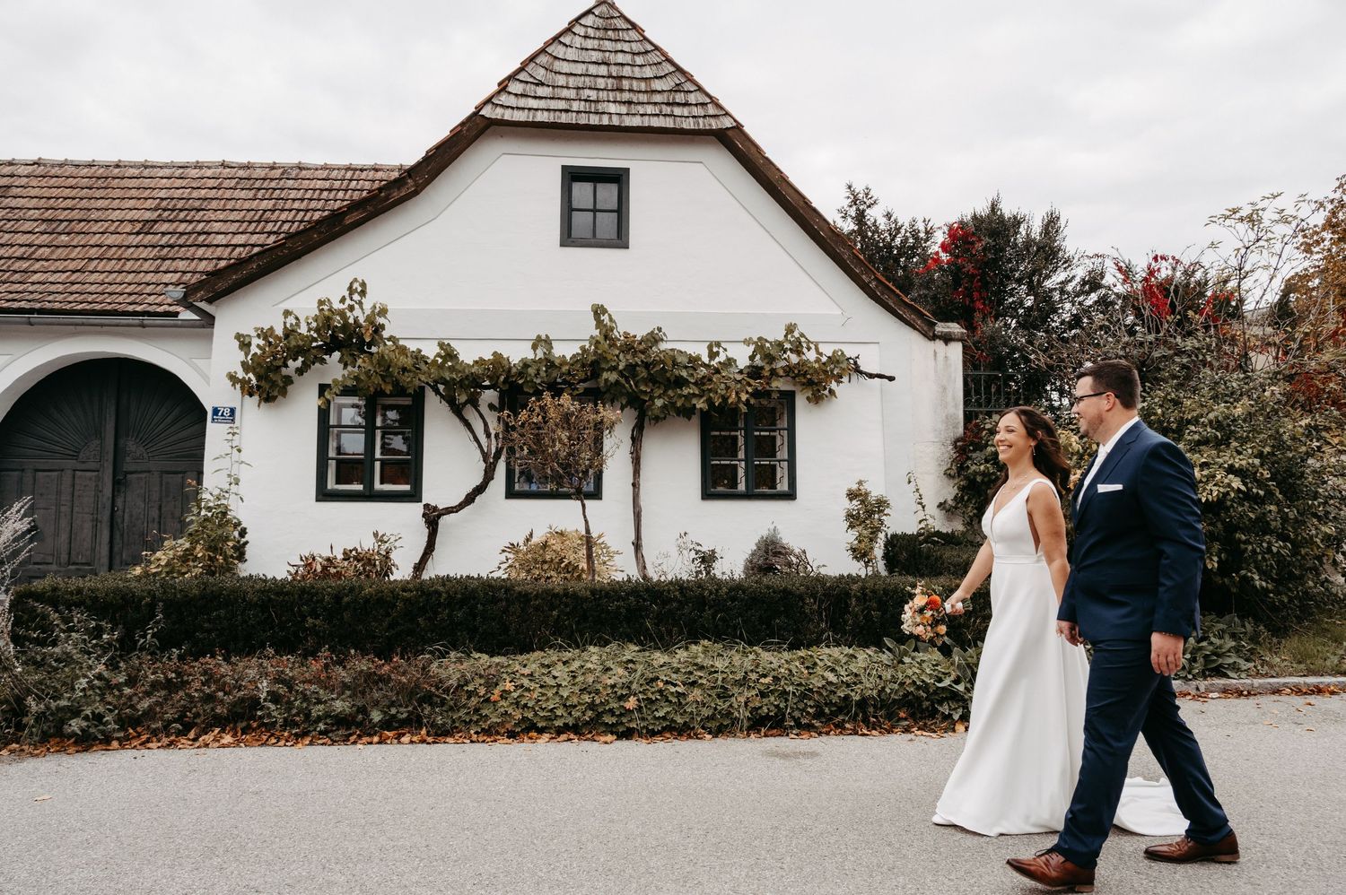Bride and groom walking hand in hand past a charming white farmhouse with climbing roses during autumn wedding.
