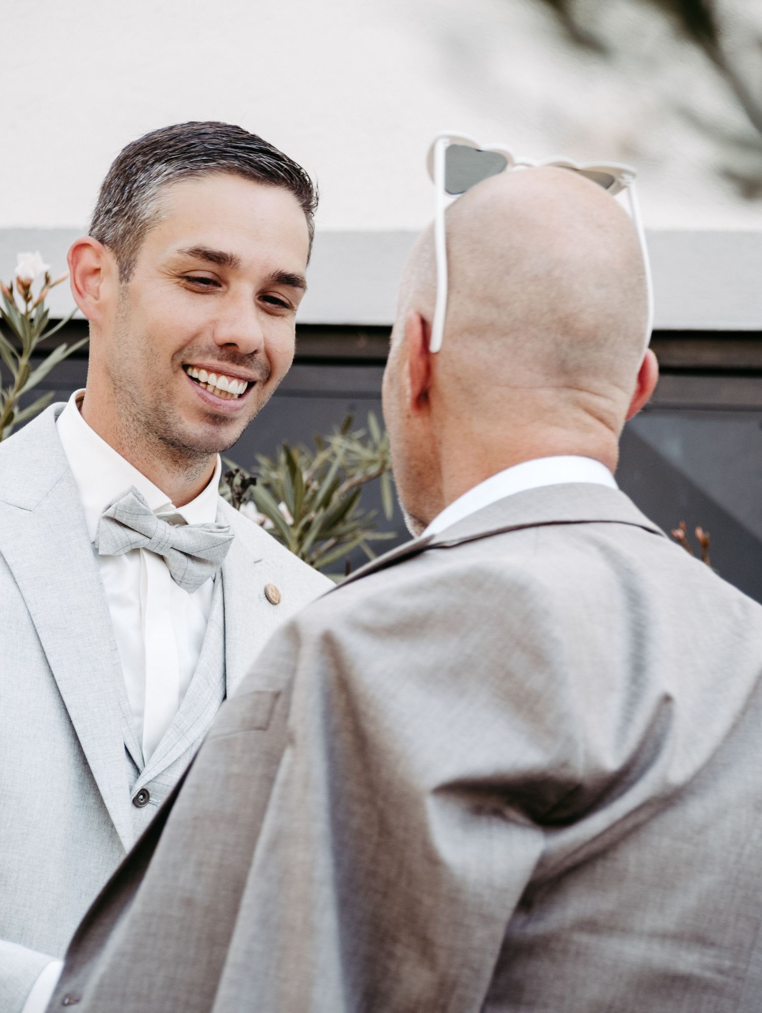 Two grooms in gray suits share a joyful moment during their wedding ceremony outdoors.