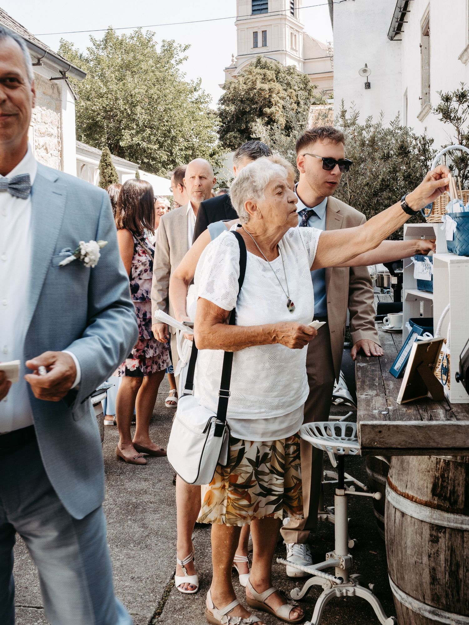 Elderly woman at outdoor wedding reception near wooden barrel, surrounded by guests in formal attire.