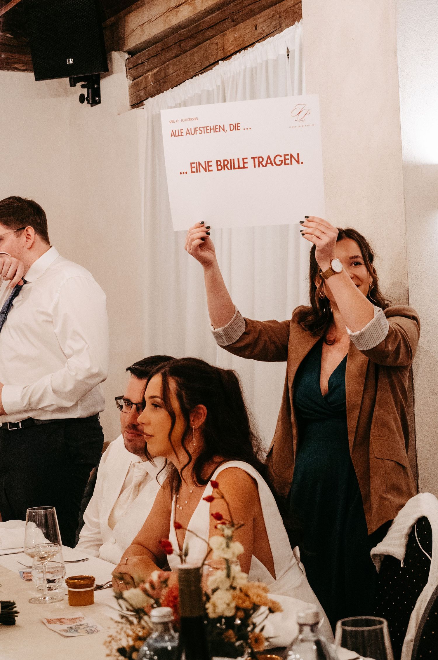 Wedding guest holds sign reading EINE BRILLE TRAGEN during fun reception party game.
