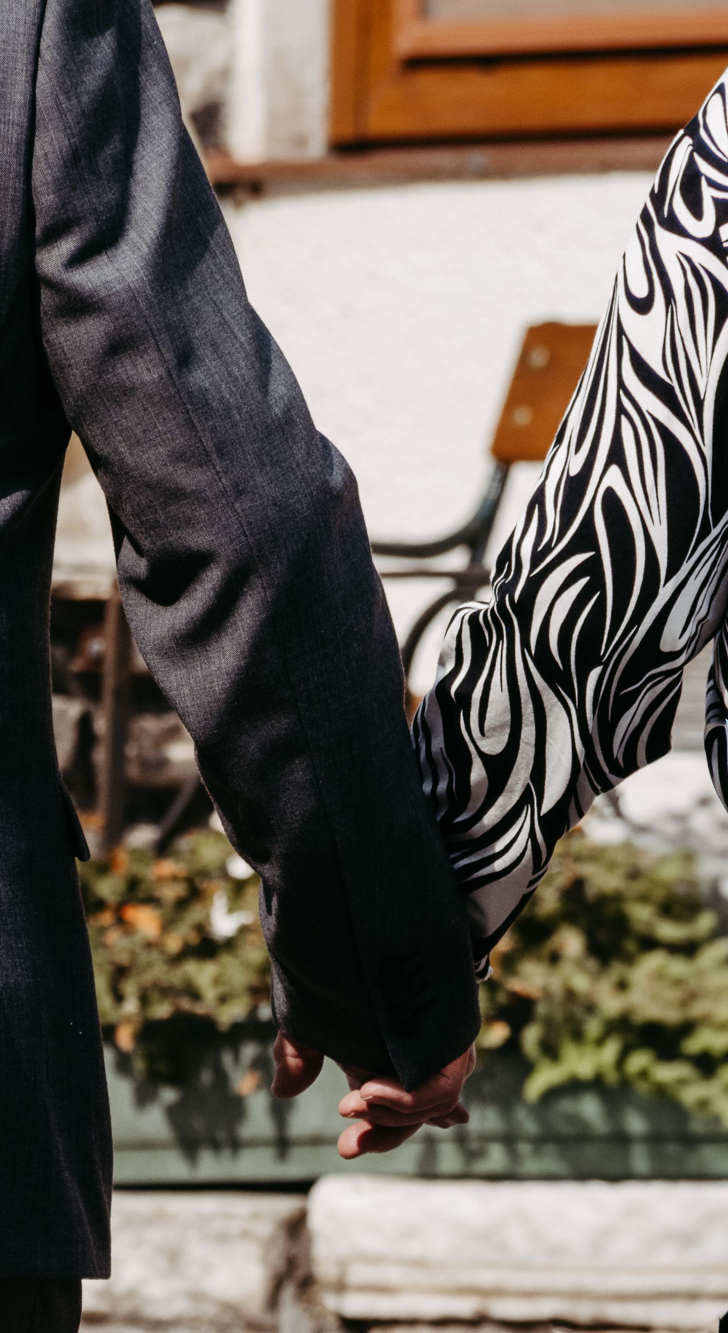Couple holding hands, man in dark suit and woman in black and white patterned dress, standing outdoors near greenery.