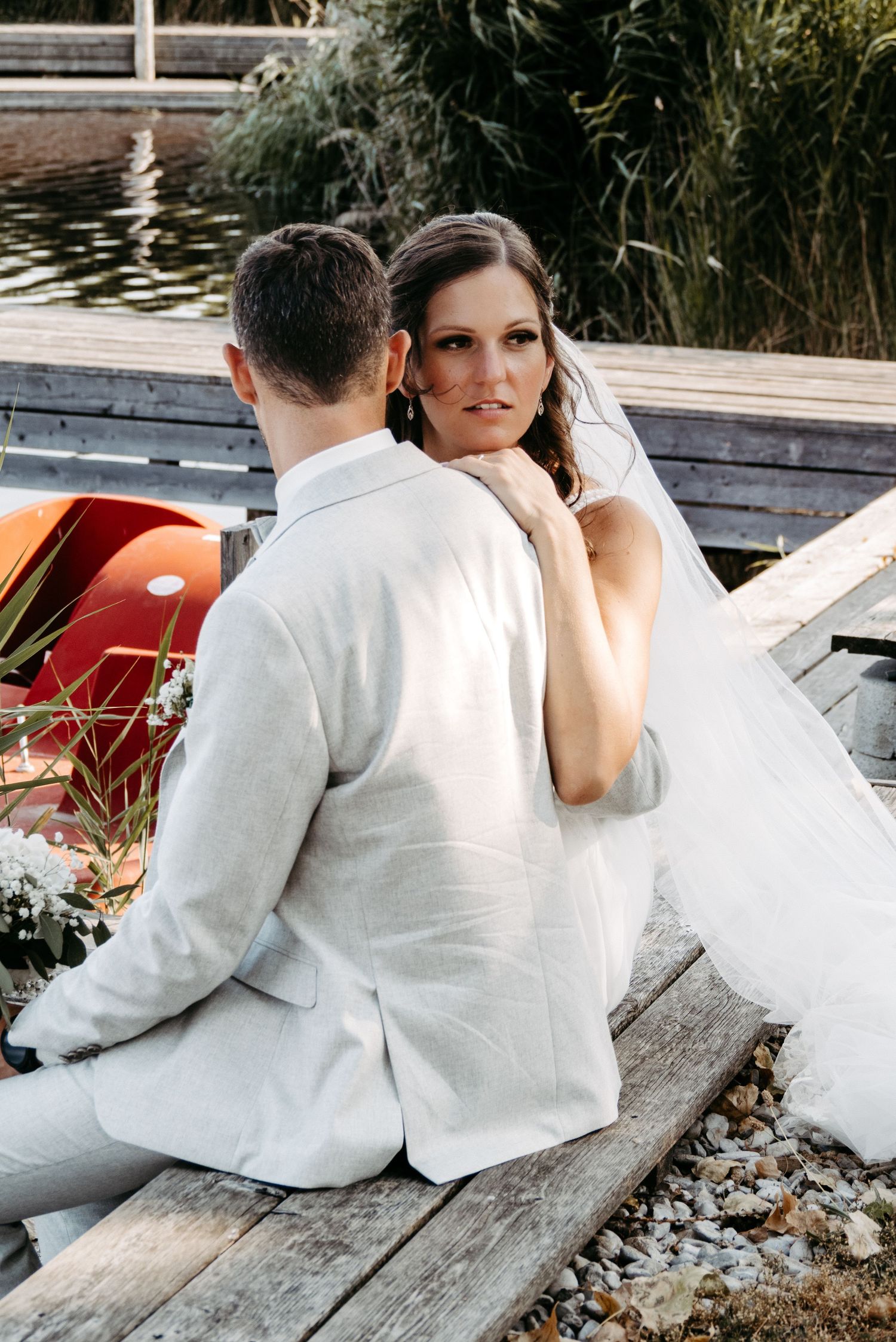 Bride resting her chin on groom's shoulder while sitting on a wooden dock near a lake on their wedding day.