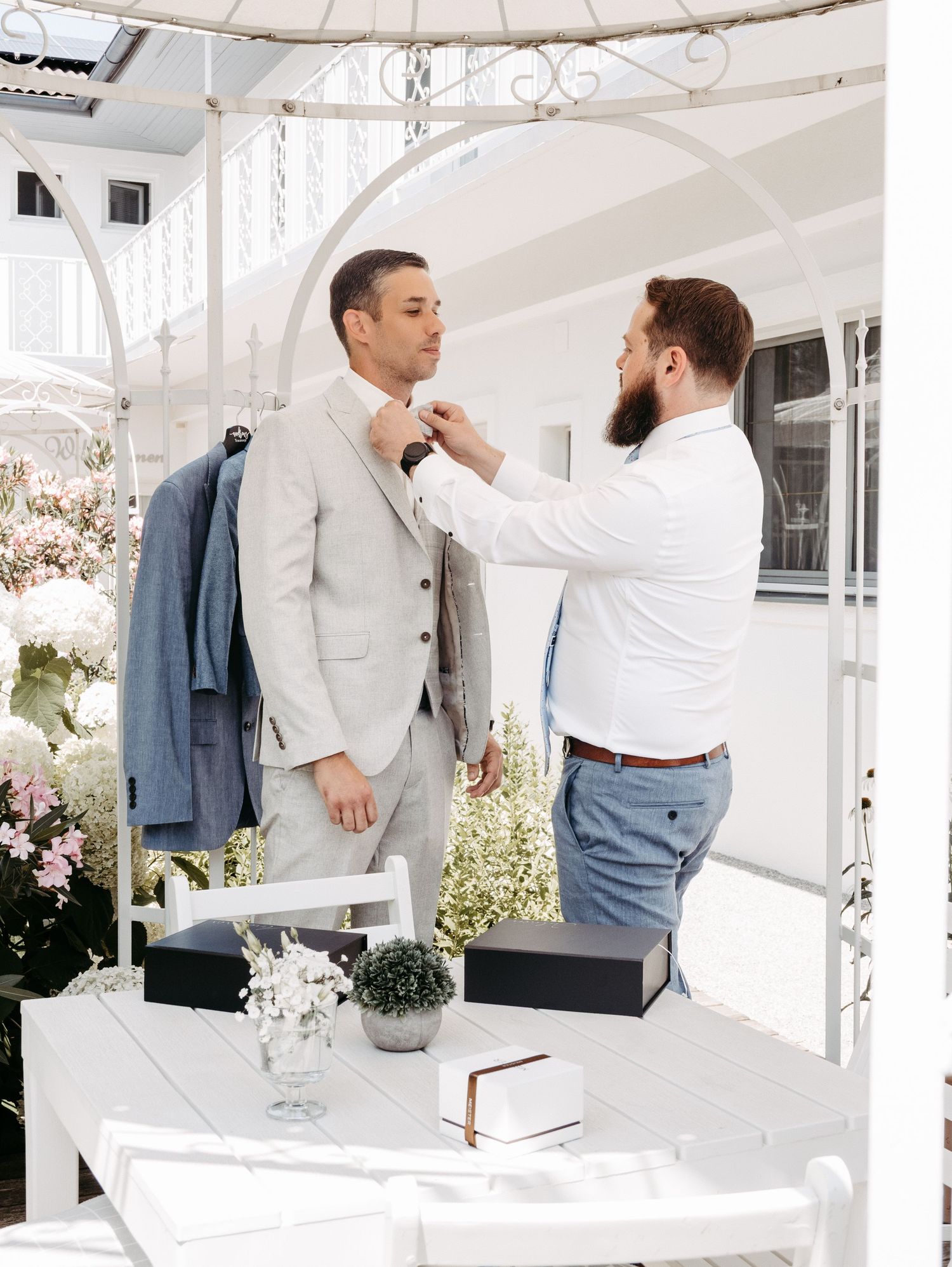 A bearded man adjusts the tie of another man in a light gray suit inside an elegant white floral boutique.