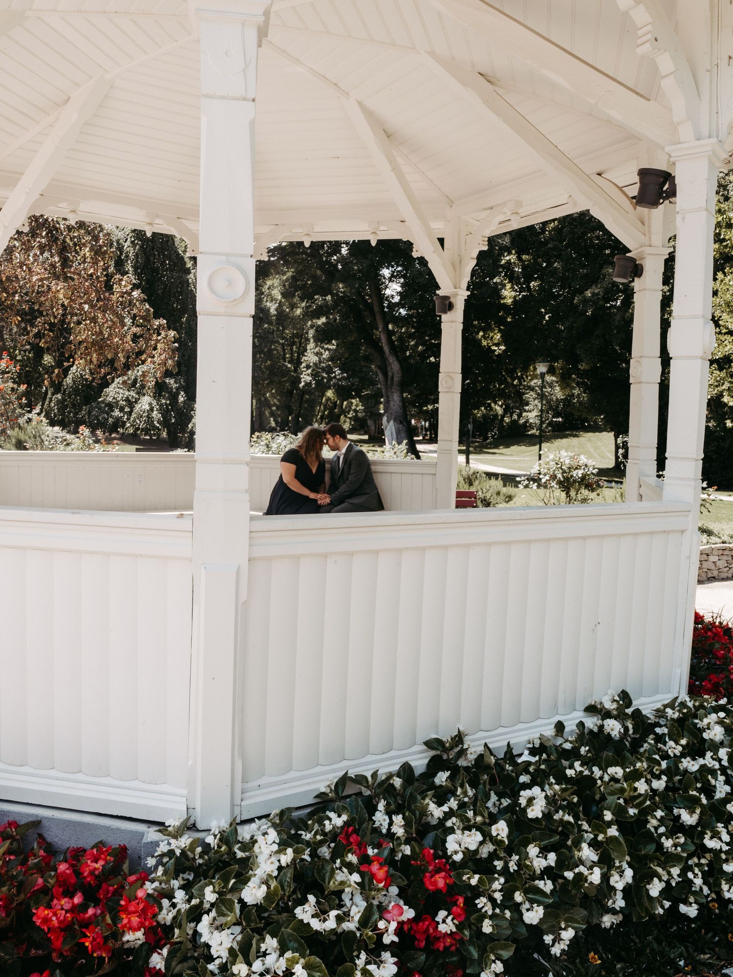 Couple sharing a romantic moment in a white gazebo surrounded by red and white flowers in a park.